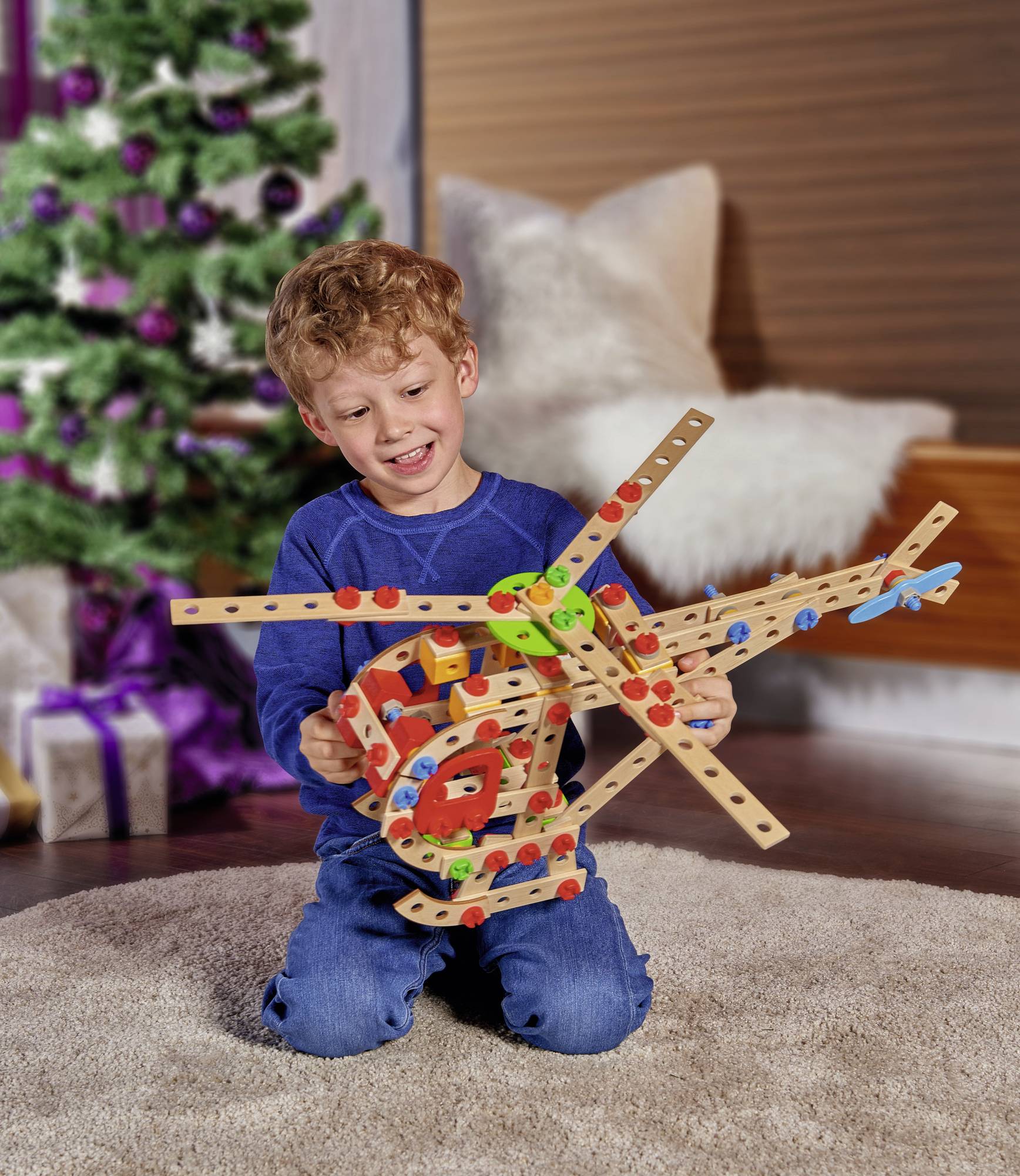 Een kind zit op een tapijt en bouwt een kleurrijk speelgoedmodel van houten stukken met schroeven. Op de achtergrond staat een versierde kerstboom.