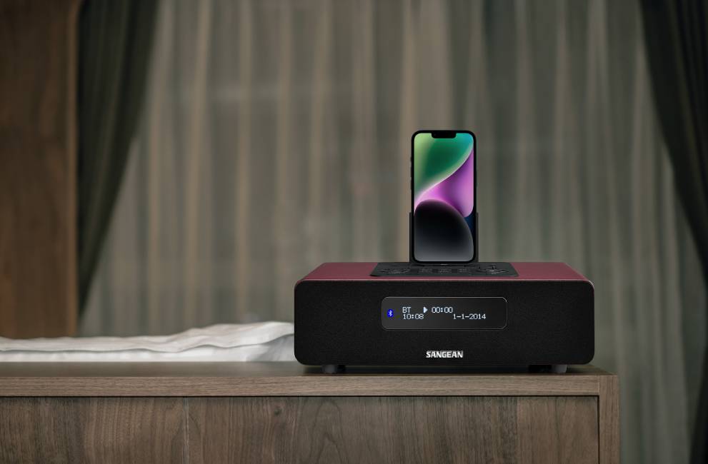 Een smartphone staat op een rode soundbox op een houten tafel, met donkere gordijnen te zien op de achtergrond.