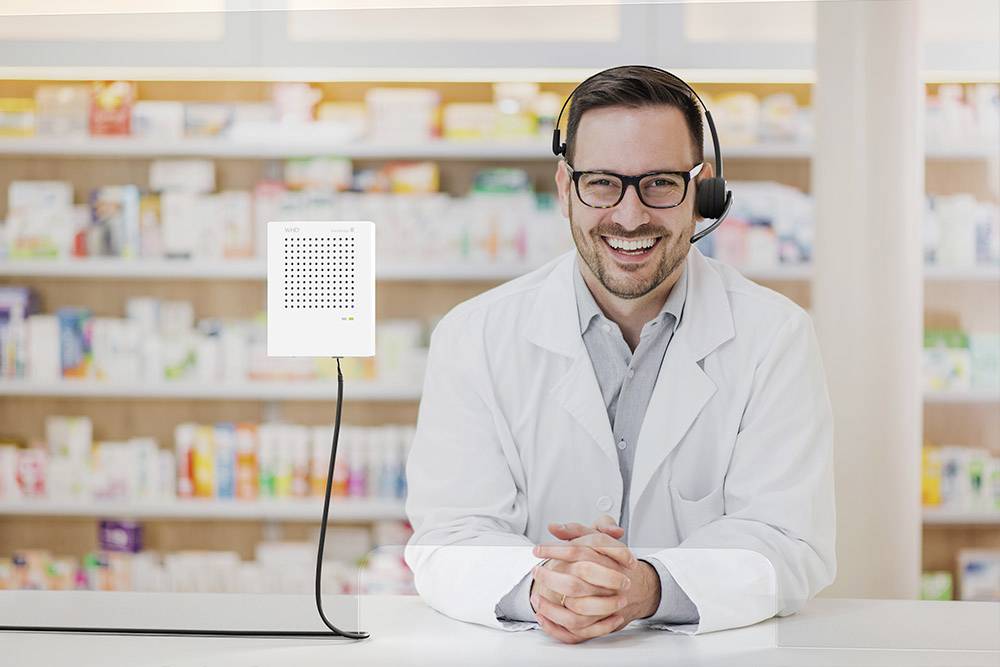 Een glimlachende apotheker met een headset staat achter de balie in een apotheek. Op de achtergrond zijn schappen met medicijnen zichtbaar.