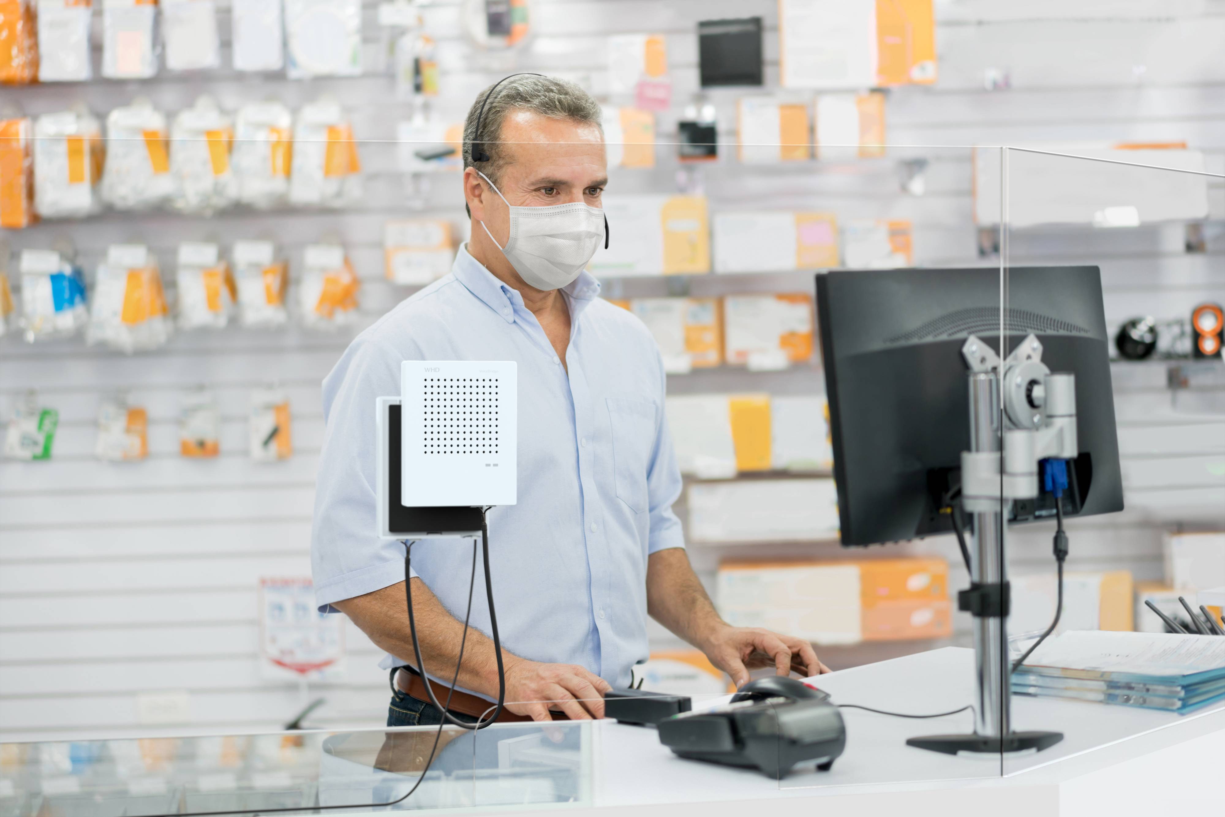Een man met een masker werkt op een computer in een winkel. Op de achtergrond zijn verschillende elektronische artikelen zichtbaar.