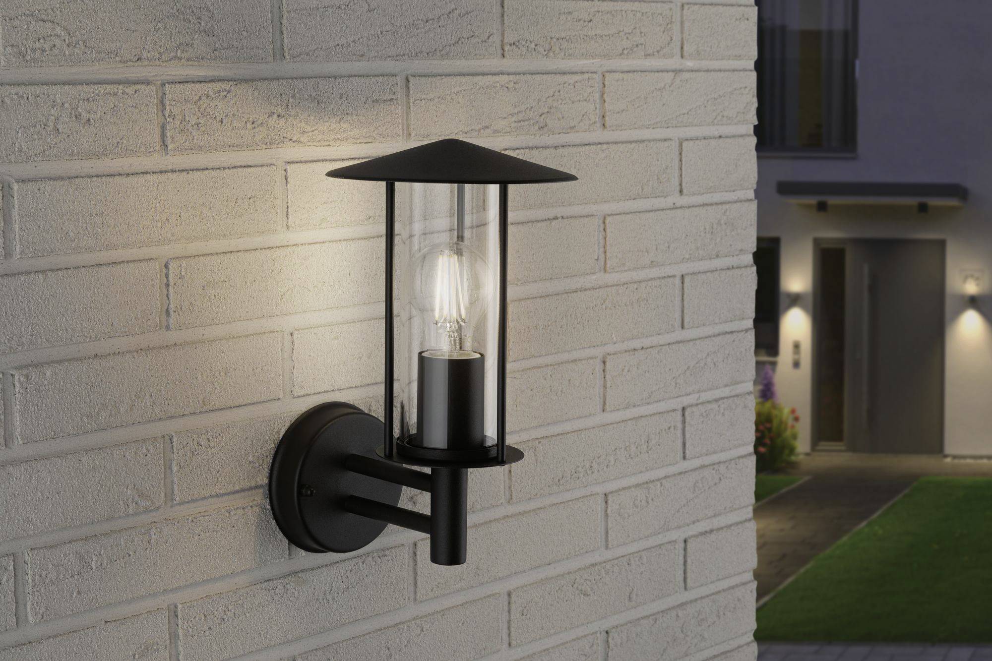 Een buitenwandlamp in klassiek design schijnt tegen een bakstenen muur. Op de achtergrond een wazig huis bij nacht.