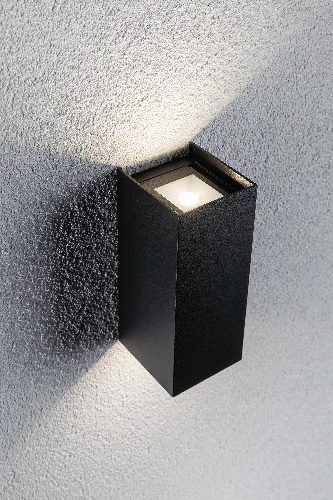Wandlamp met een zwart, rechthoekig behuizing, verlicht zowel naar boven als naar beneden, creëert lichtpatronen op een ruwe, grijze wandoppervlakte.
