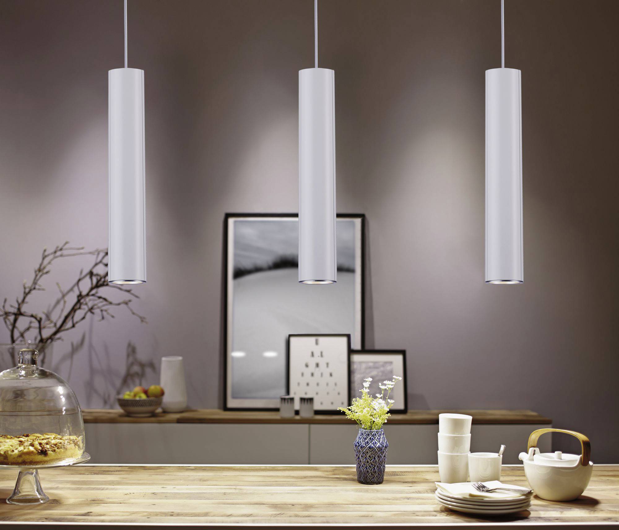 Keuken met hangende lampen, een houten tafel met servies en bloemen, achtergrond met kunstwerken. Gezellige sfeer.