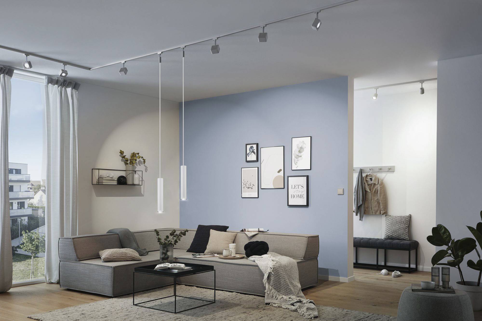 Een modern woonkamer met een blauwe accentmuur. Een grijze bank met kussens en dekens, met daarboven fotolijsten. Heldere verlichting.