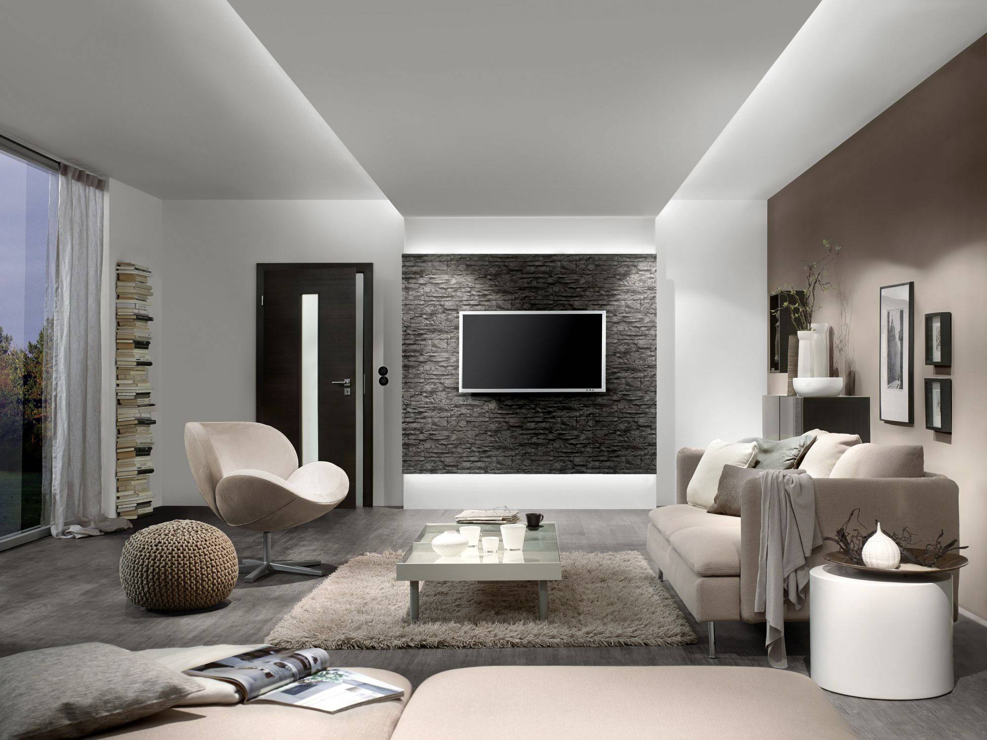 Modern ingericht woonkamer met lichte meubels, een flatscreen-tv aan een stenen muur en grote ramen aan de linkerkant.