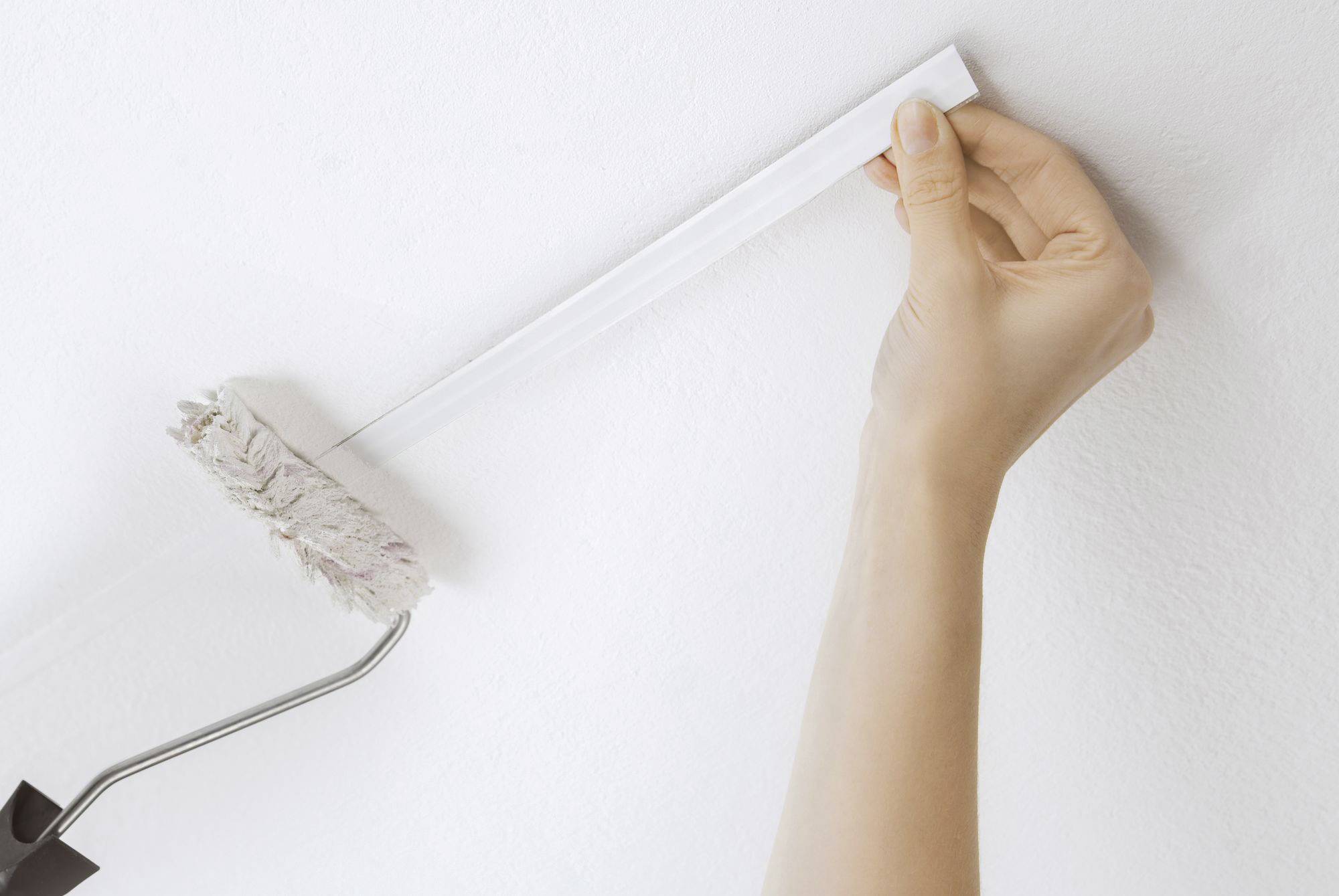 Een hand verwijdert voorzichtig schilderstape van een witte muur, terwijl een verfroller met witte verf erop rust.