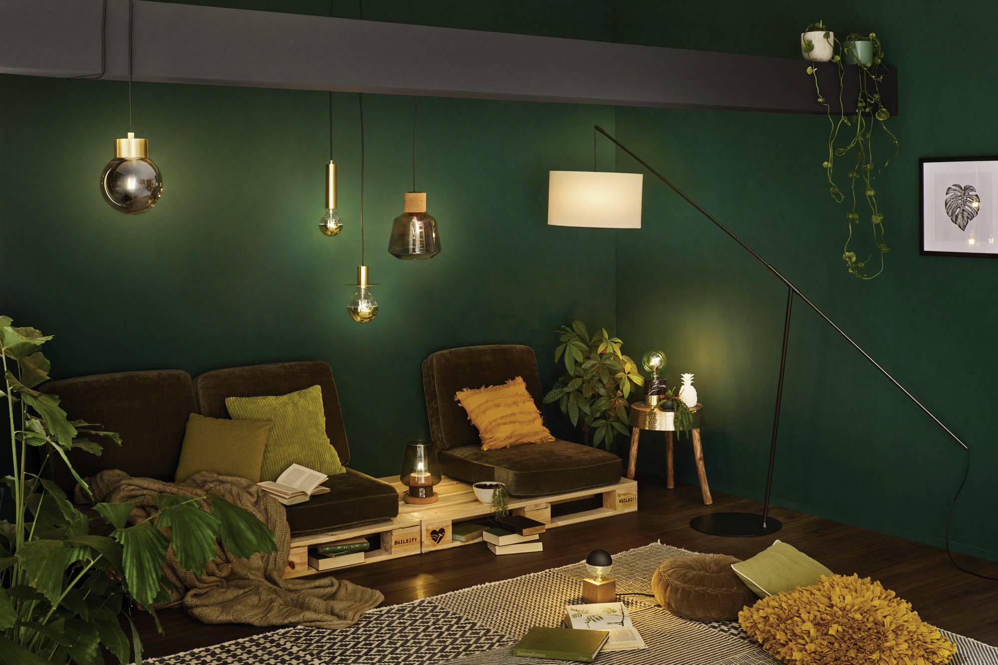 Gezellige woonkamer met groene muren, bank van houten pallets met bruine kussens, planten, boeken, lampen en een vloerkleed.
