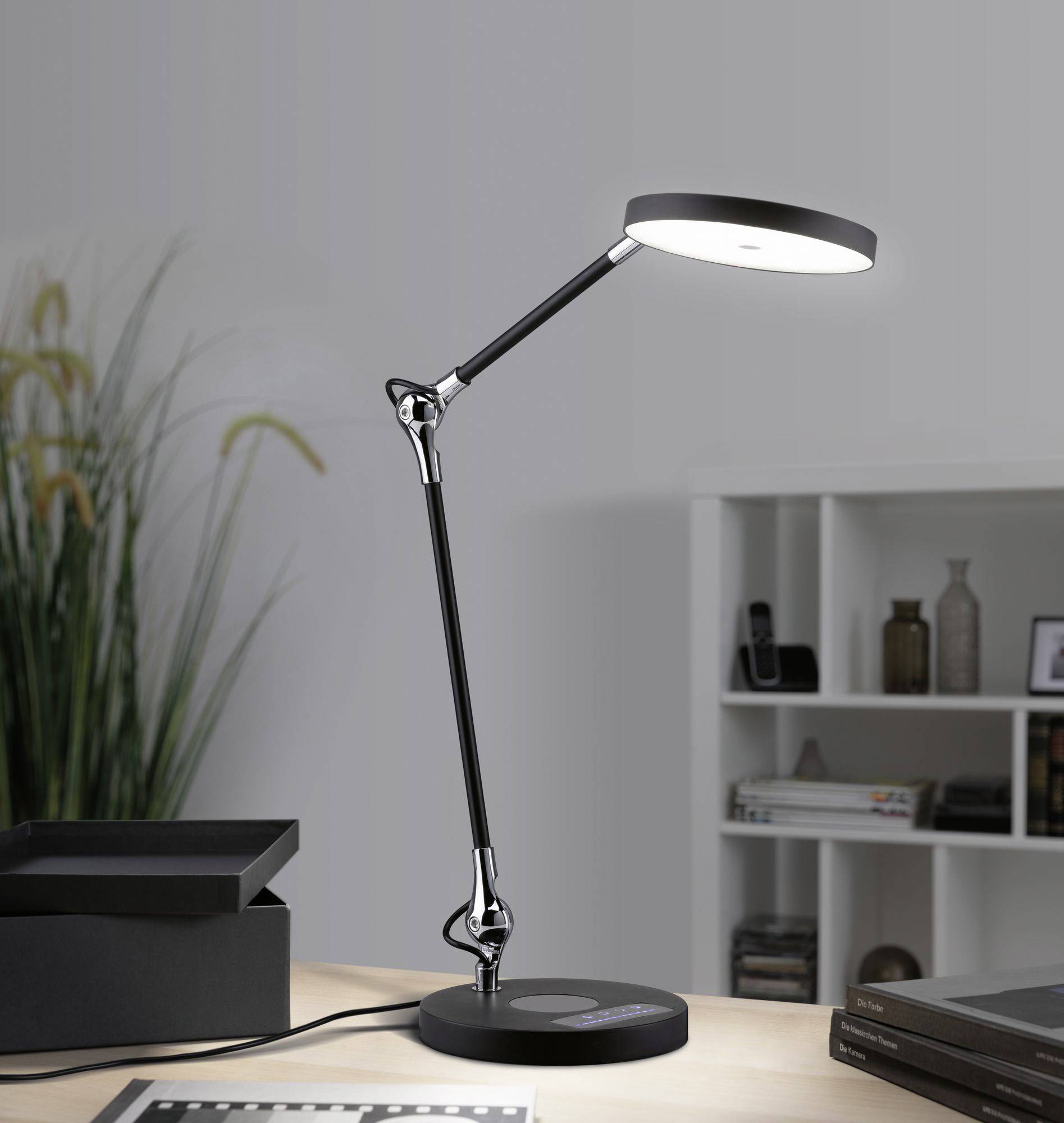 Een moderne bureaulamp verlicht een werkruimte. Op de achtergrond zijn een boekenkast en een kamerplant zichtbaar.
