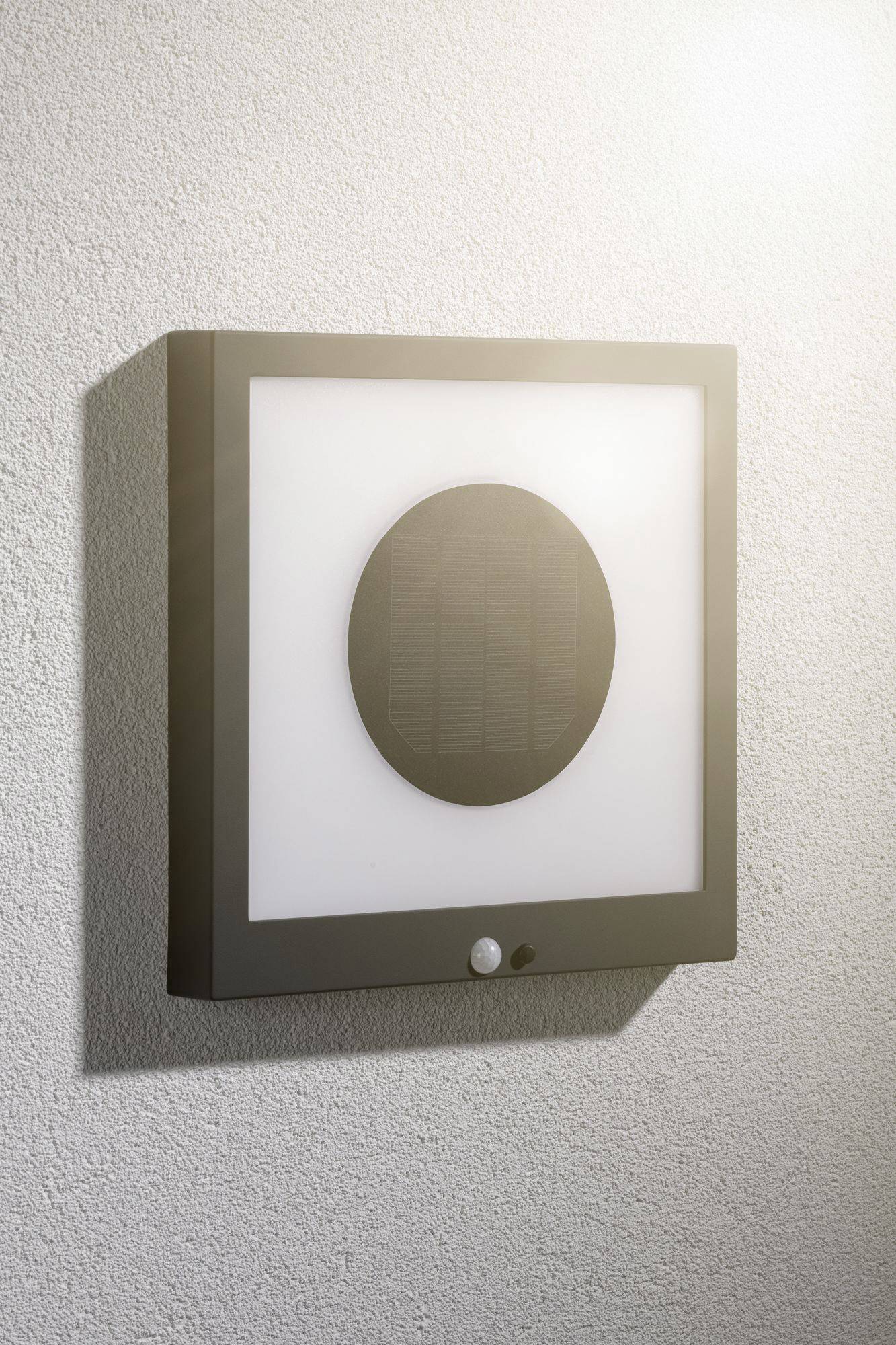 Vierkante buitenlamp aan een muur, met een rond zonnepaneel in het midden en een bewegingssensor eronder. Ideaal voor energiezuinige verlichting.