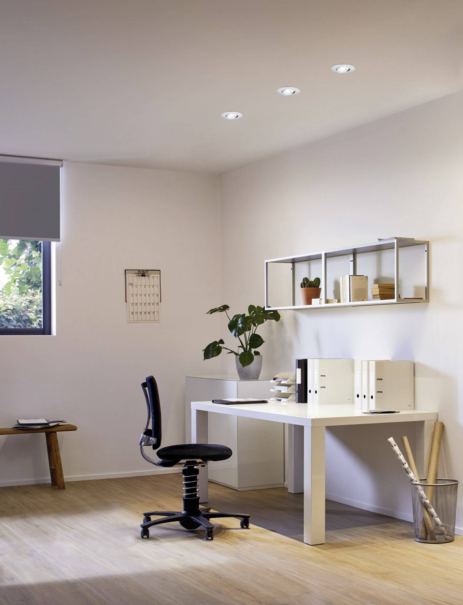 Een minimalistisch thuiskantoor met een witte bureau, zwarte stoel, wandplanken, planten en mappen, verlicht door plafondspots.