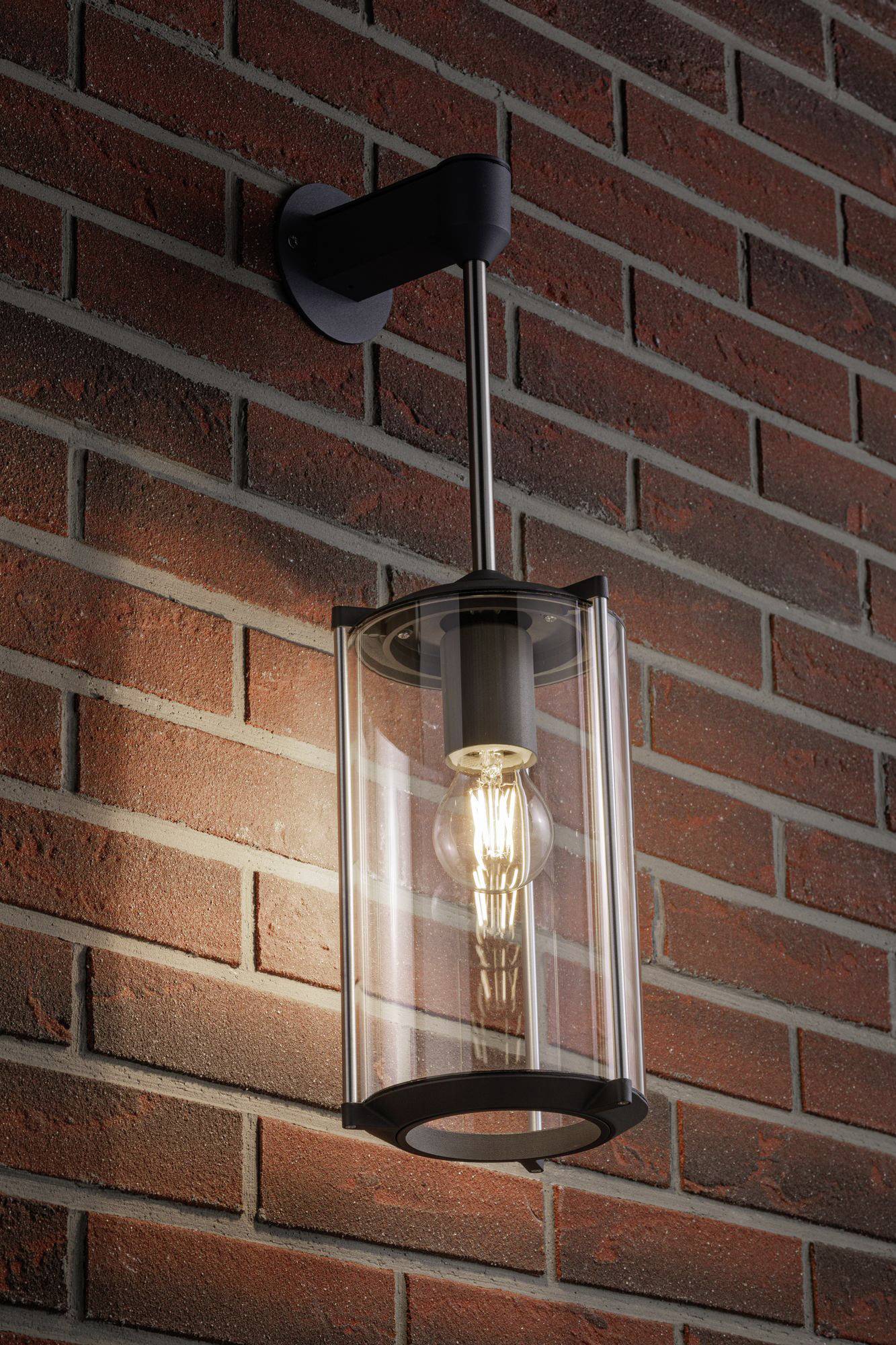 Buitenwandlamp met helder glas en zwart frame, gemonteerd aan een bakstenen muur. De lamp is ingeschakeld.