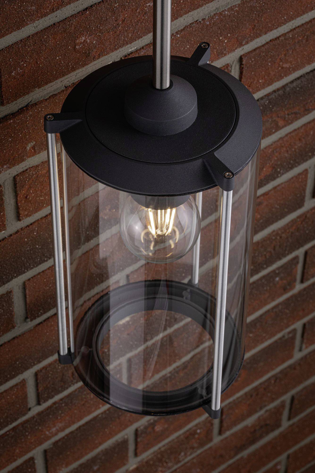 Een zwarte buitenlamp met een helder glazen behuizing, die aan een bakstenen muur hangt. De gloeilamp binnenin is zichtbaar.
