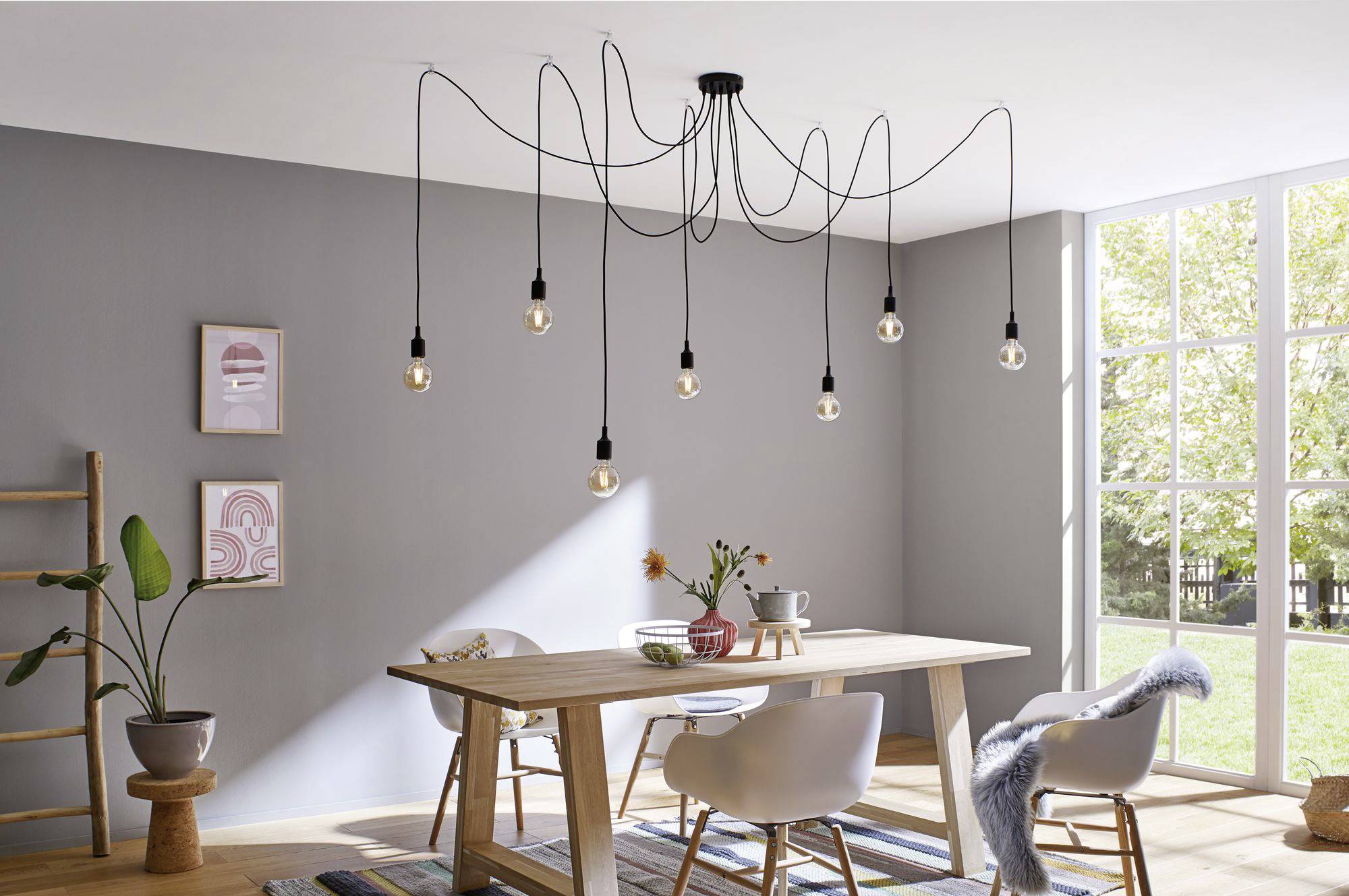 Modern eetkamergedeelte met een lange houten tafel, witte stoelen en een opvallende plafondlamp met meerdere loshangende gloeilampen.