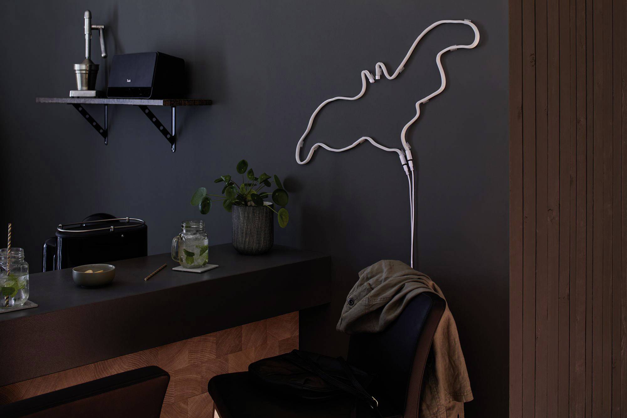 Een stijlvol café met donker interieur, een bar met twee drankjes en planten erop. Aan de muur hangt een neonlicht in de vorm van een vleermuis.