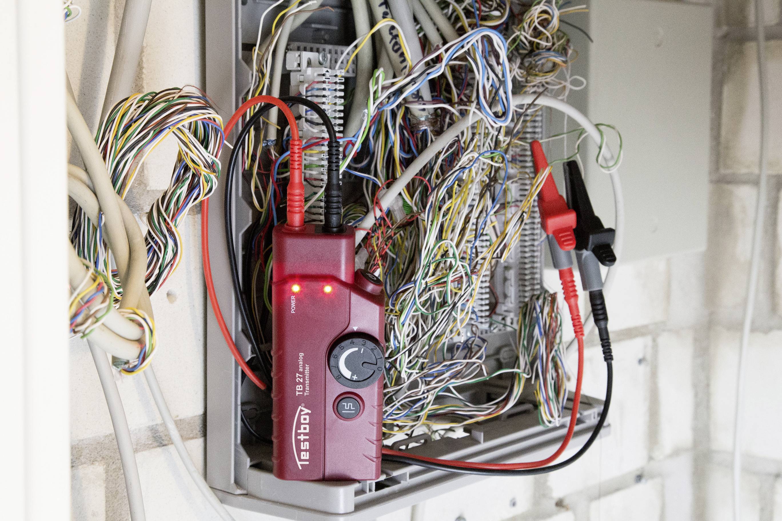 Een rood diagnostisch apparaat met oplichtende LED's is aangesloten op een complex netwerk van gekleurde kabels in een verdeelkast.