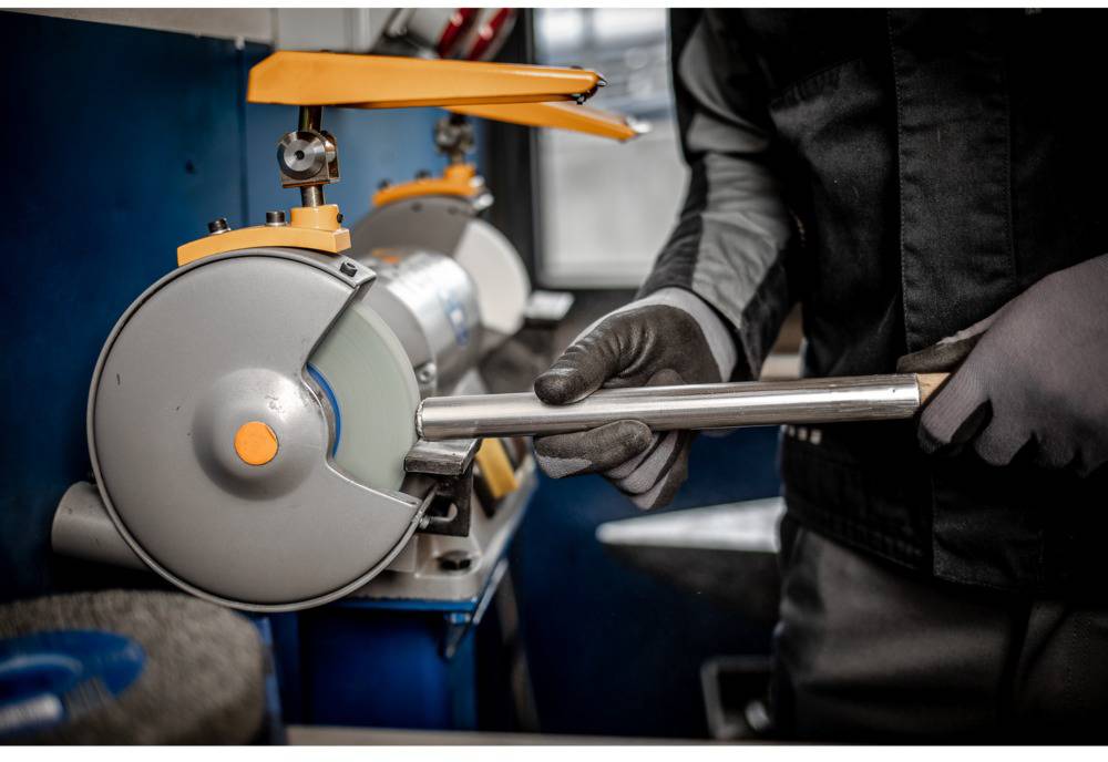 Een persoon slijpt een metalen gereedschap met een slijpmachine in een werkplaats. De persoon draagt veiligheidskleding en handschoenen.