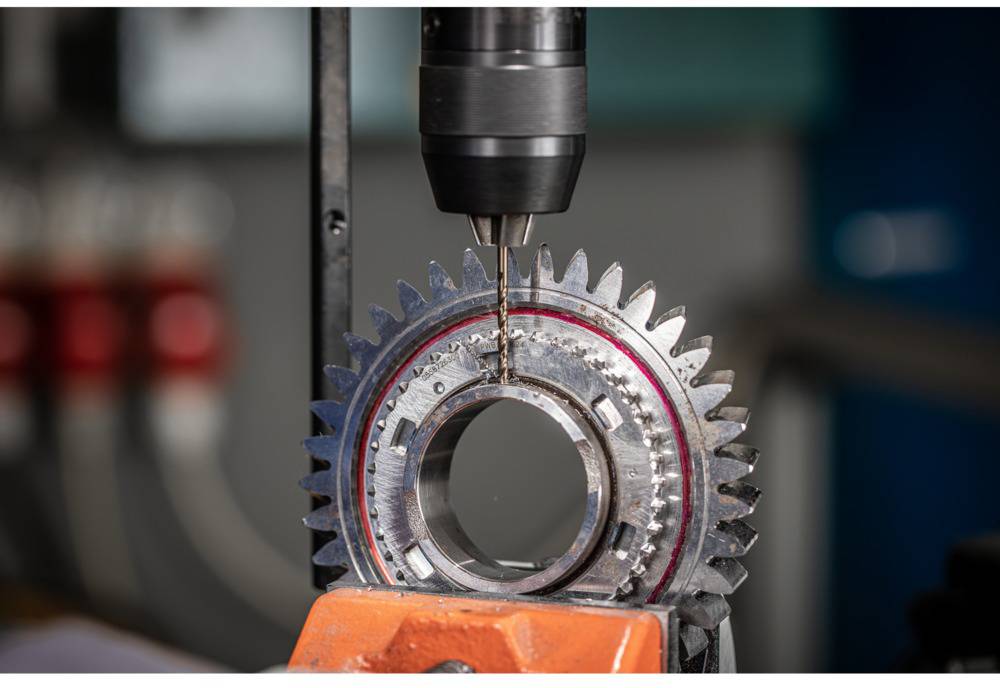 Een boor bewerkt een tandwiel van metaal. De scène toont een fabricageproces in een werkplaatsomgeving.