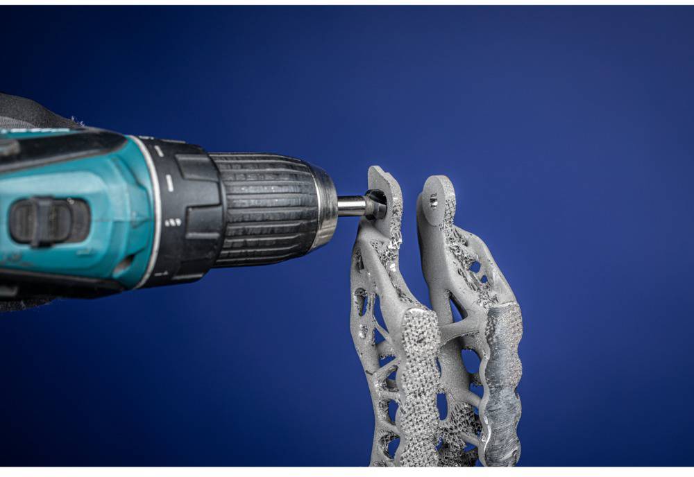 Een boor bewerkt een grijs, complex 3D-geprint onderdeel voor een blauwe achtergrond.