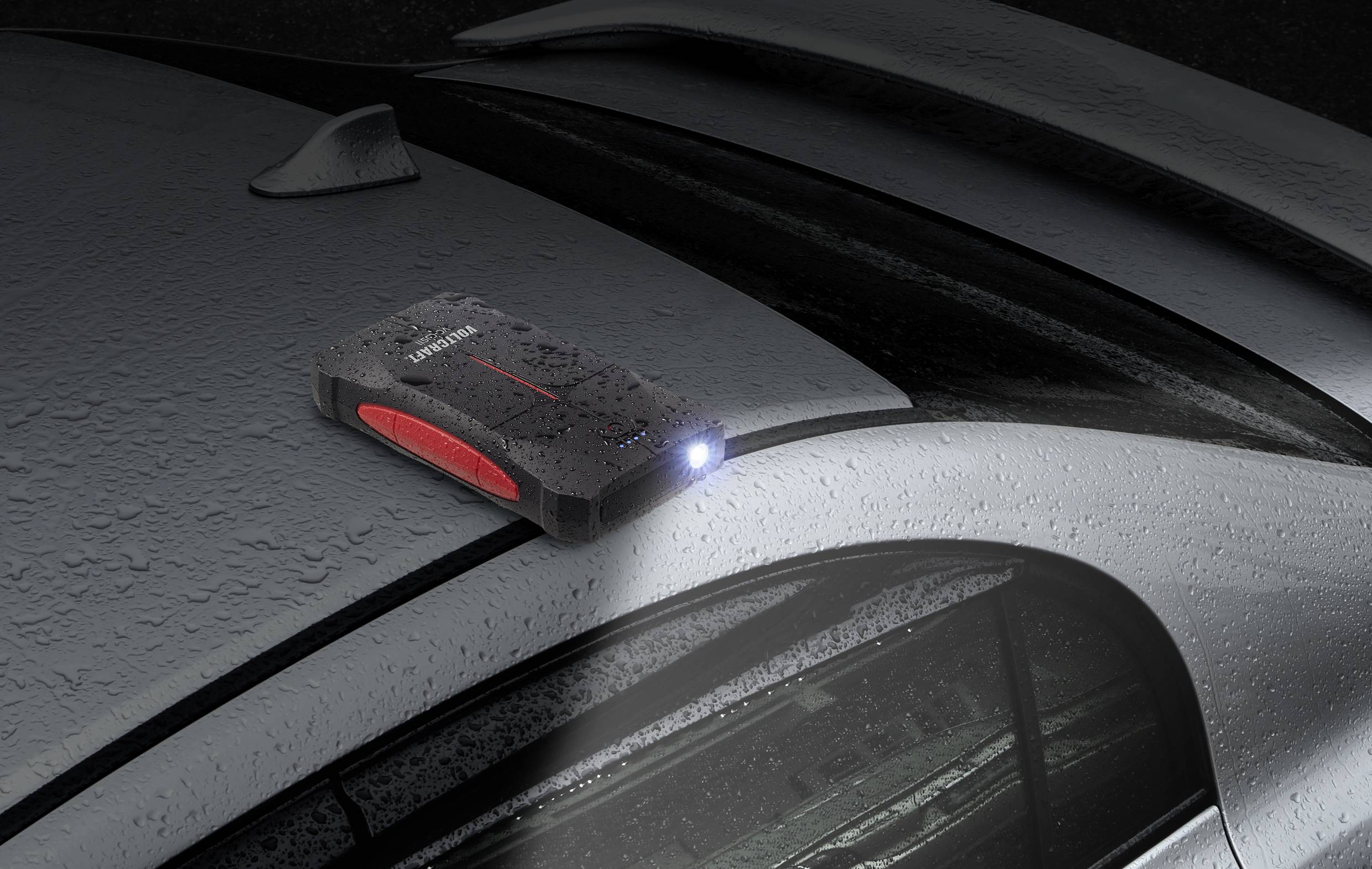 Een draagbare starthulpaccu met ingeschakelde zaklamp ligt op een nat autodak.