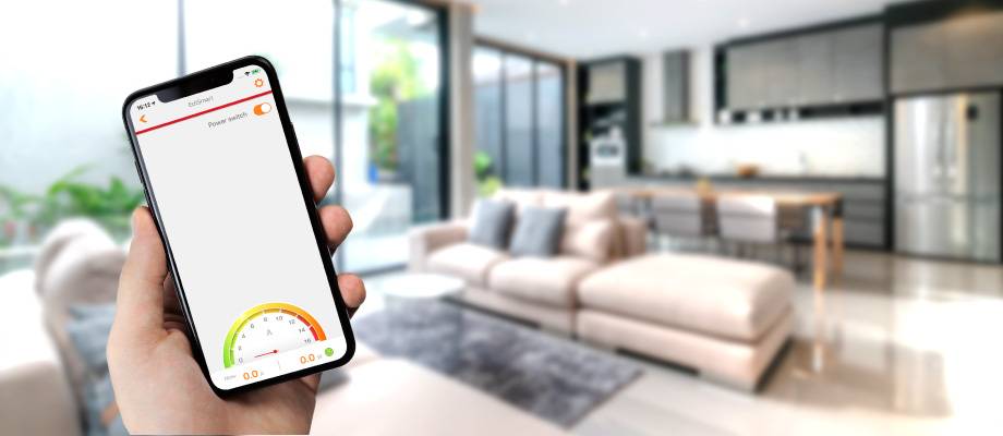 Een persoon houdt een smartphone vast met een app voor energieverbruik. Op de achtergrond een moderne woonkamer en keuken.