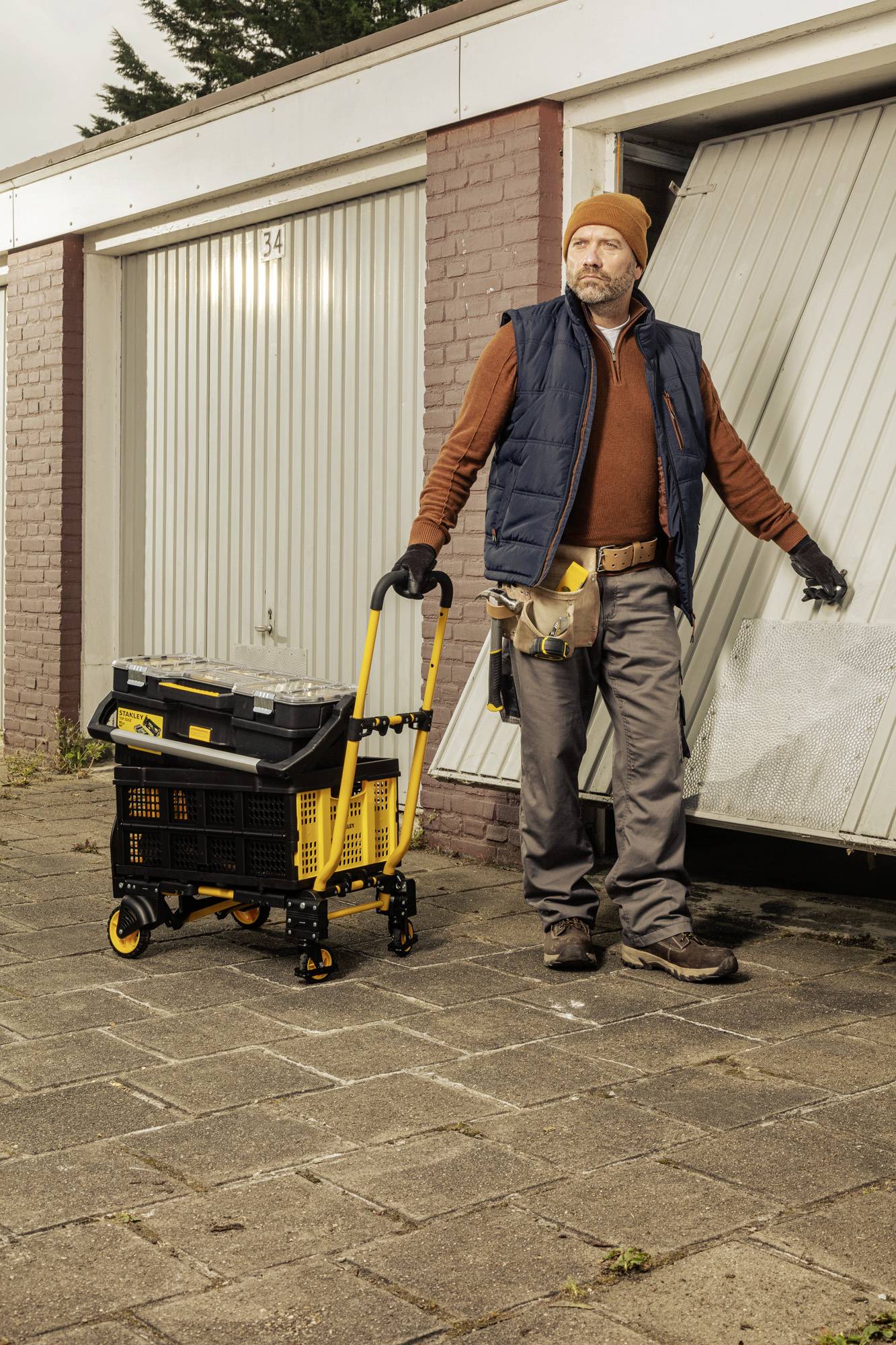 Een persoon in werkkleding opent een garagedeur en houdt een gele gereedschapswagen vast. De scène oogt werkgericht en praktisch.