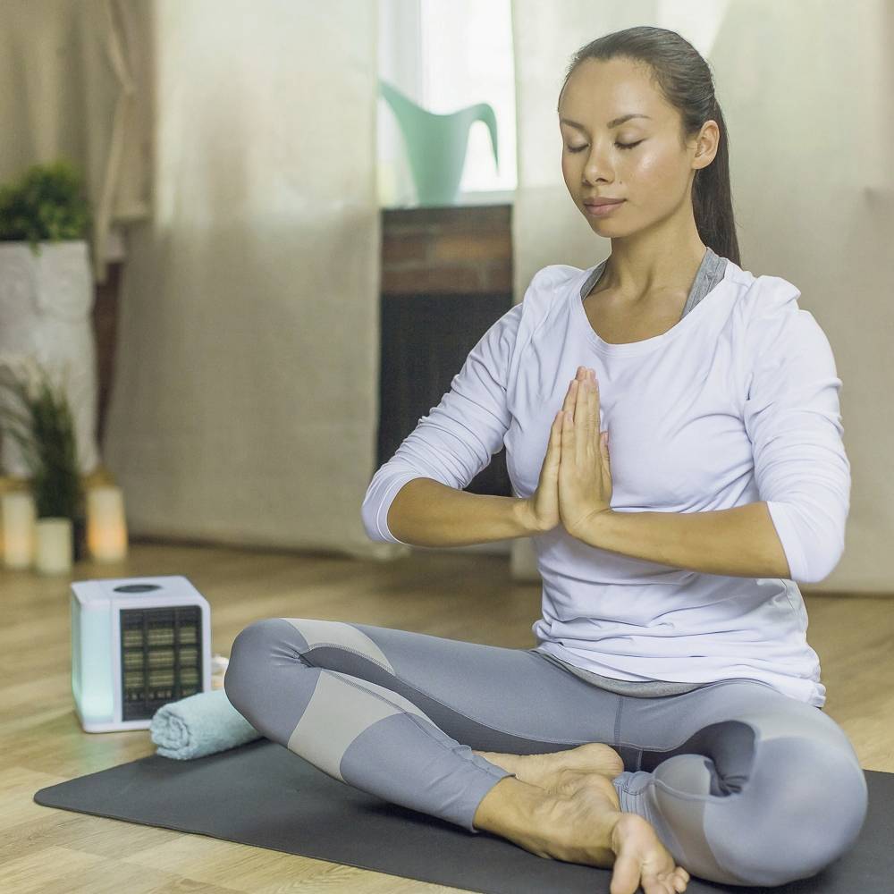 Een vrouw in meditatiehouding zit op een yogamat in een ruimte met kaarsen en planten, die rust en ontspanning uitstraalt.