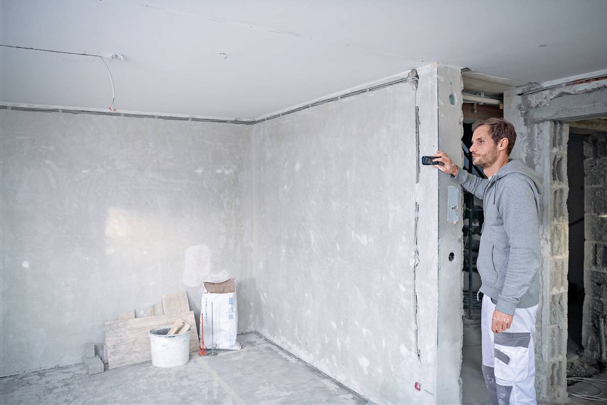 Een persoon meet de muur met een lasermeetinstrument in een onafgewerkte ruimte. Bouw- of renovatiewerkzaamheden zijn aan de gang.