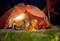 Twee personen liggen 's nachts in een verlichte tent op een weide, omringd door kampeeruitrusting onder een sterrenheldere hemel.
