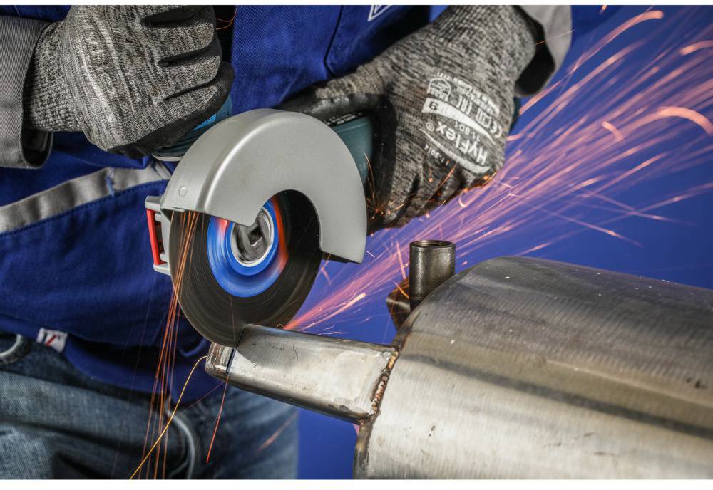 Een persoon slijpt een metalen stuk met een haakse slijper, vonken spatten. Draagt beschermende kleding, een blauwe vest en grijze handschoenen.
