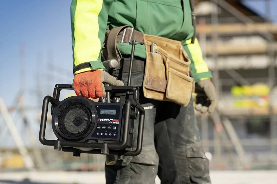 Een bouwvakker in werkkleding houdt een draagbare radio vast op een bouwterrein. Op de achtergrond zijn bouwheistellingen wazig te zien.