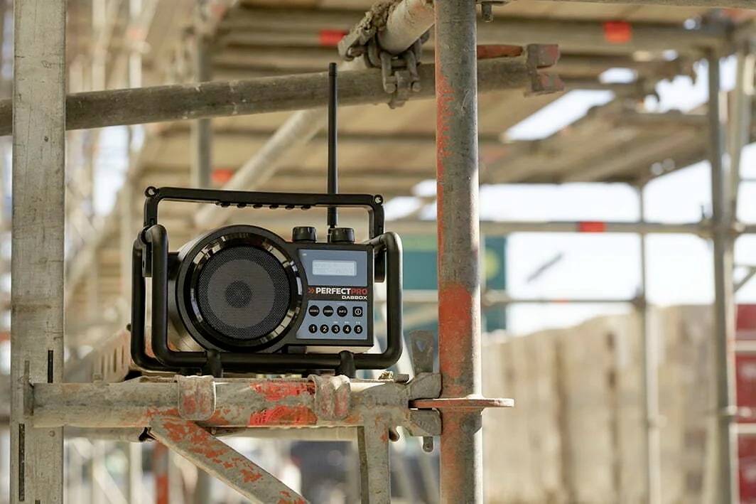 Een draagbare radio staat op een steiger. Hij is robuust gebouwd, met een antenne en verschillende bedieningsknoppen, in een bouwomgeving.