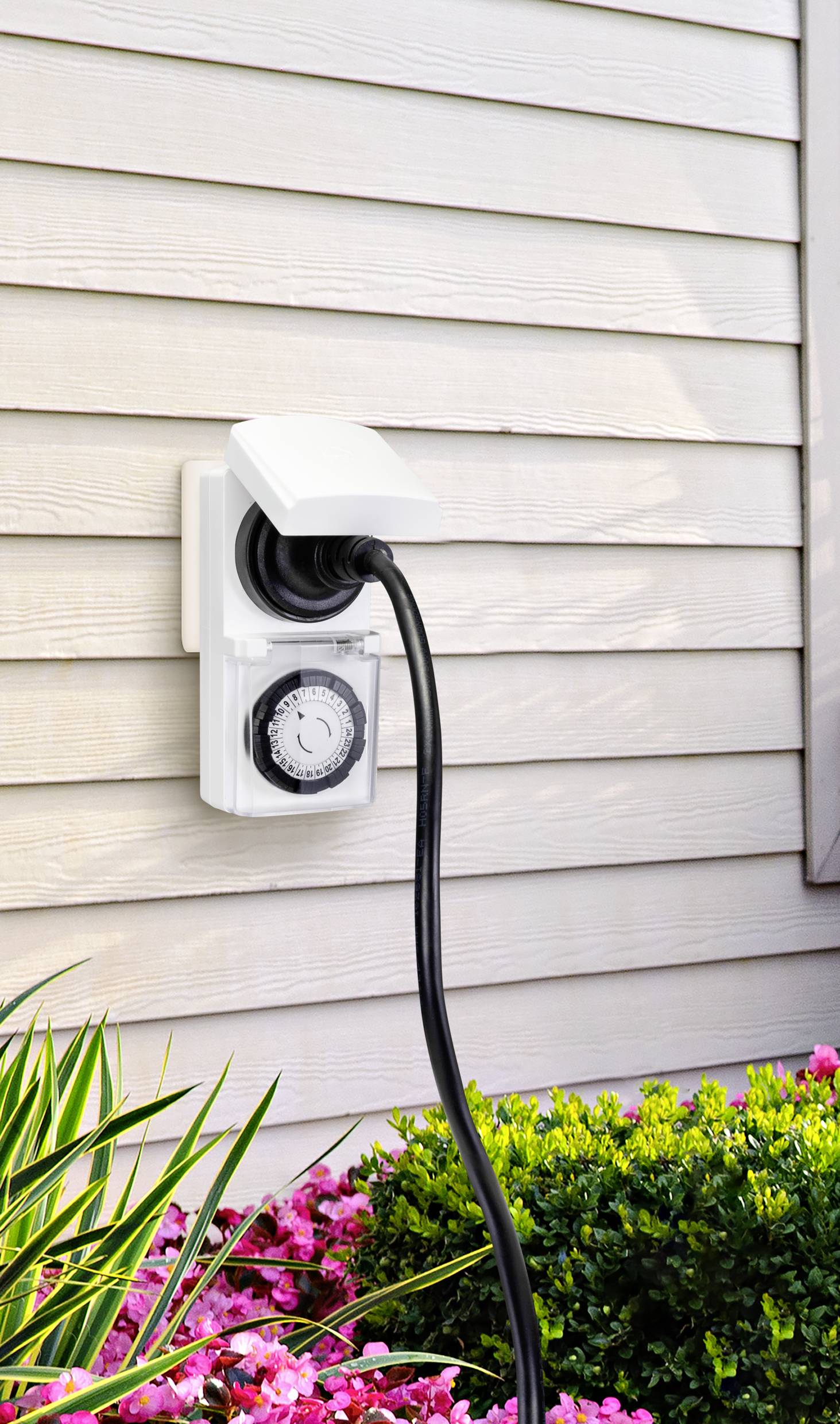 Een buitenwandcontactdoos met tijdschakelaar is aan een muur bevestigd. Een kabel is aangesloten. Op de voorgrond zijn planten zichtbaar.