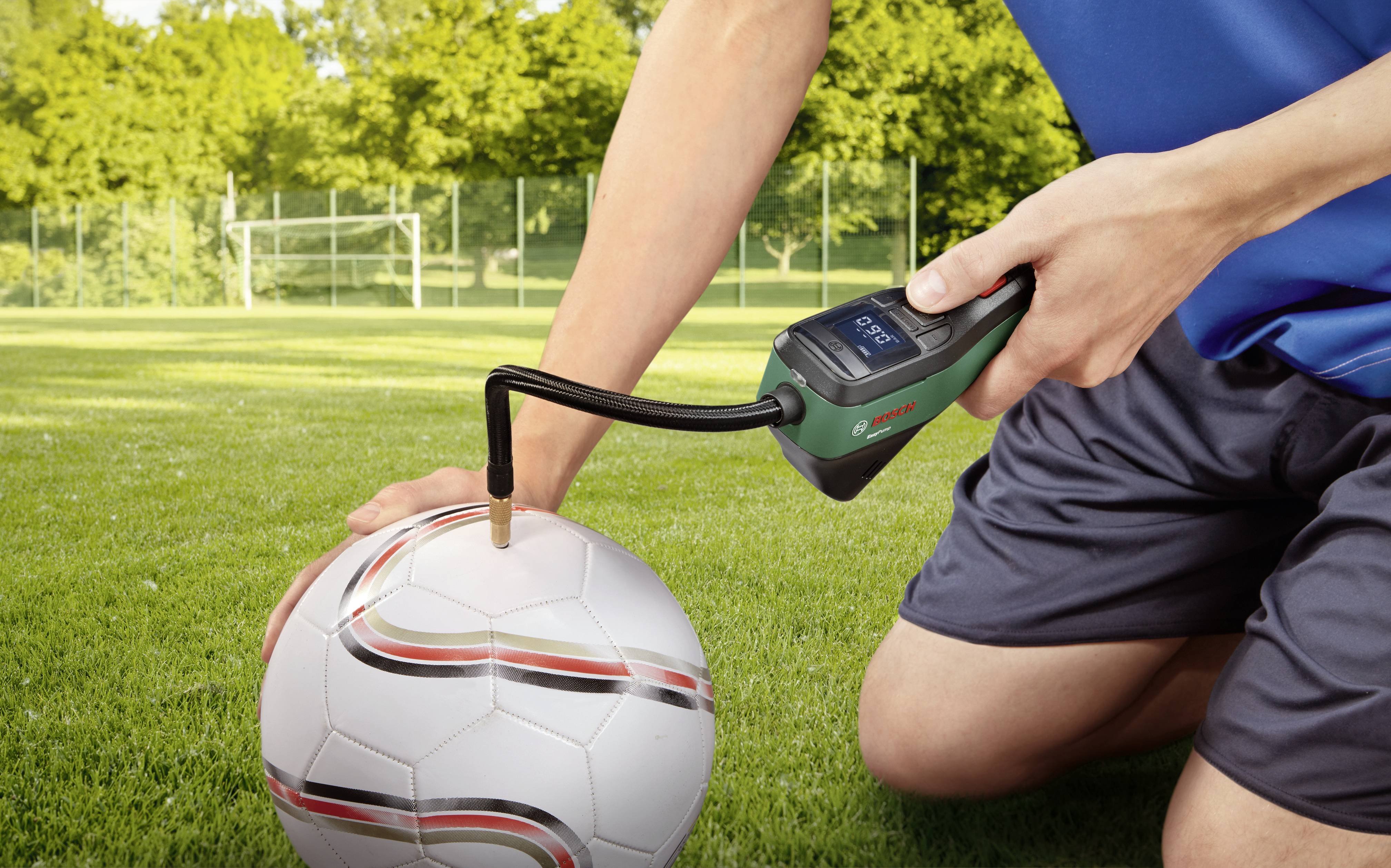 Een persoon pompt een voetbal op met een elektrische luchtpomp op een voetbalveld. Op de achtergrond is een doel zichtbaar.