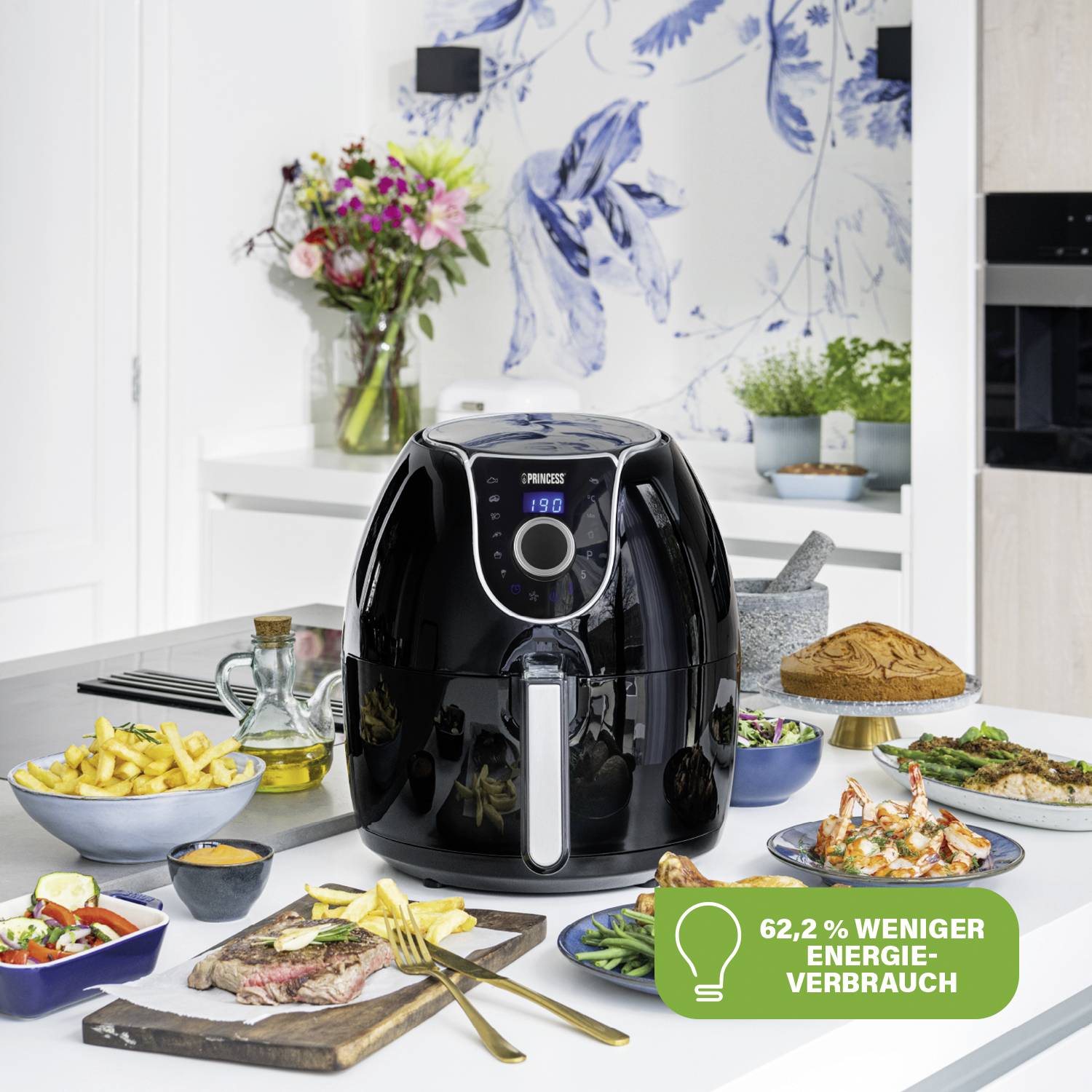 Een airfryer op een aanrecht, omringd door taart, friet en vleesgerechten. Tekst in de afbeelding: '62,2% minder energieverbruik'.