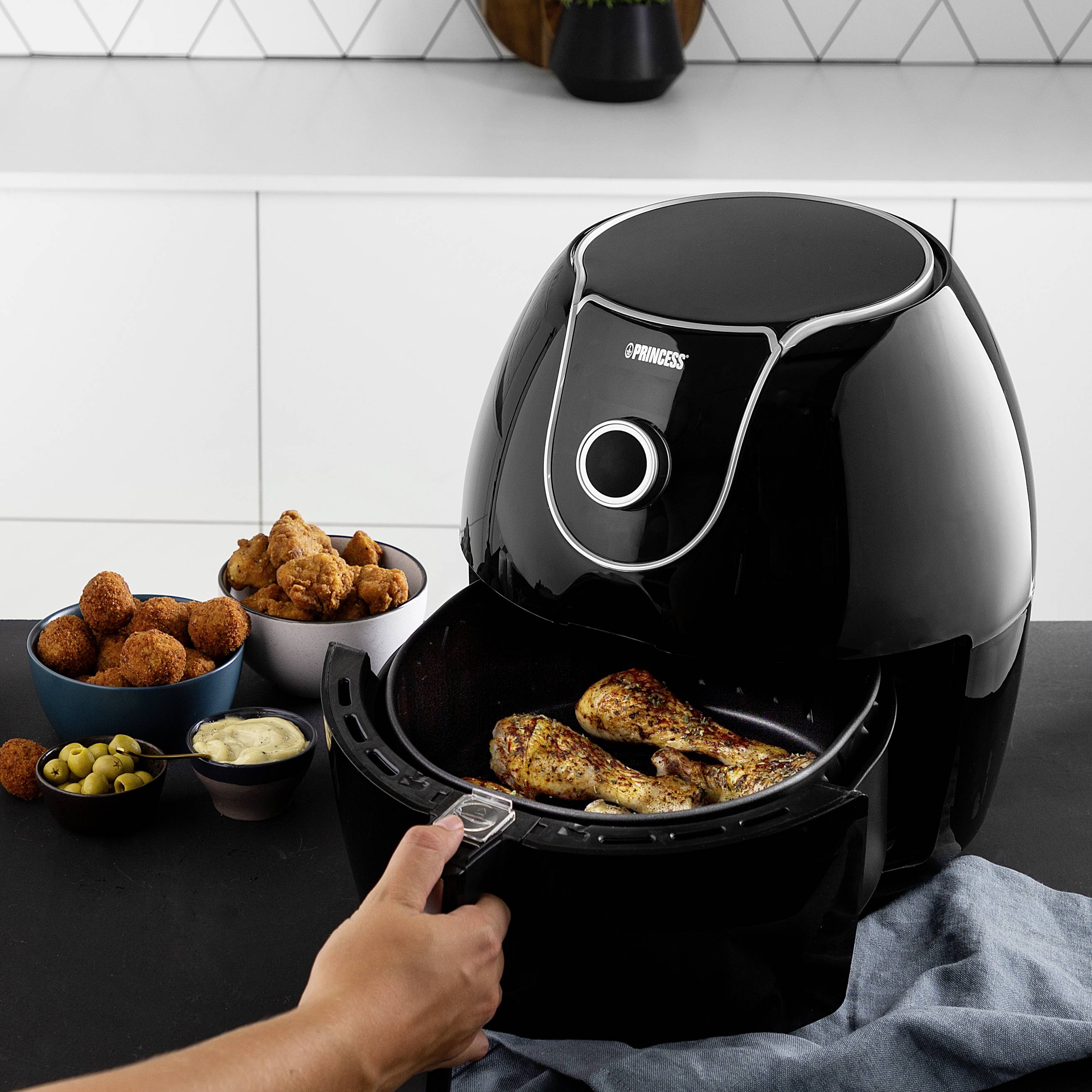 Een persoon haalt gebakken kippenstukken uit een zwarte airfryer. Op de achtergrond staan snacks en sauzen.