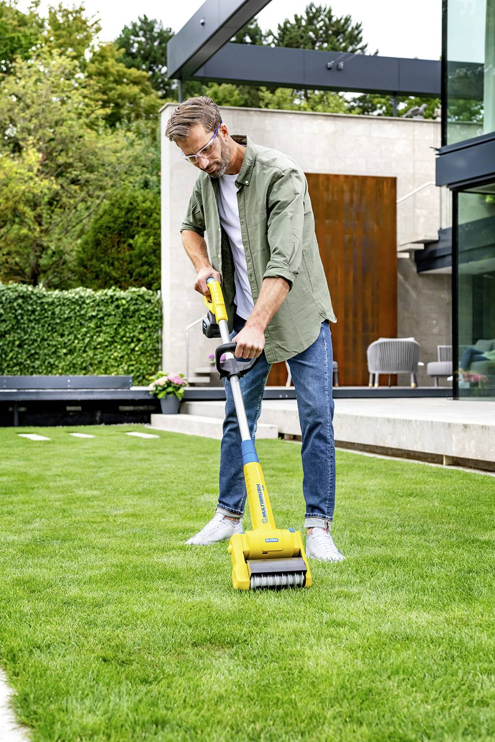 Een man maait het gazon met een gele elektrische grasmaaier in de tuin van een modern huis.