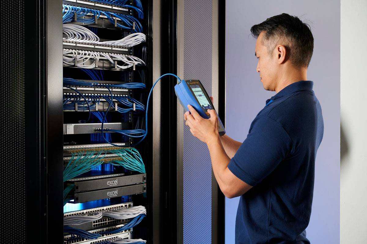 Een persoon in een blauw shirt controleert netwerkbekabeling in een serverruimte met een handapparaat.