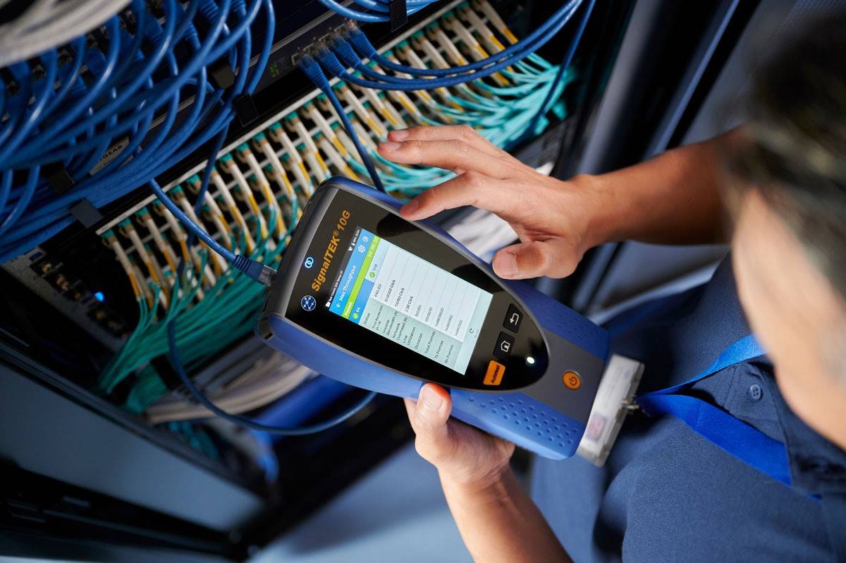 Een persoon bedient een netwerkdiagnostisch apparaat voor een serverrek met talrijke kabels om verbindingen en signaalsterkte te controleren.