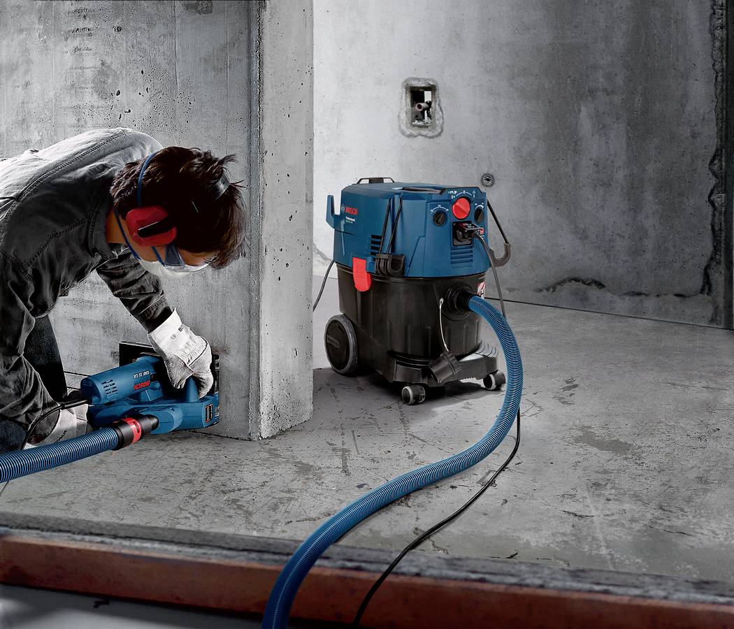 Een persoon werkt op een bouwplaats met een blauwe industriële stofzuiger en een boormachine, de betonnen muur wordt bewerkt.