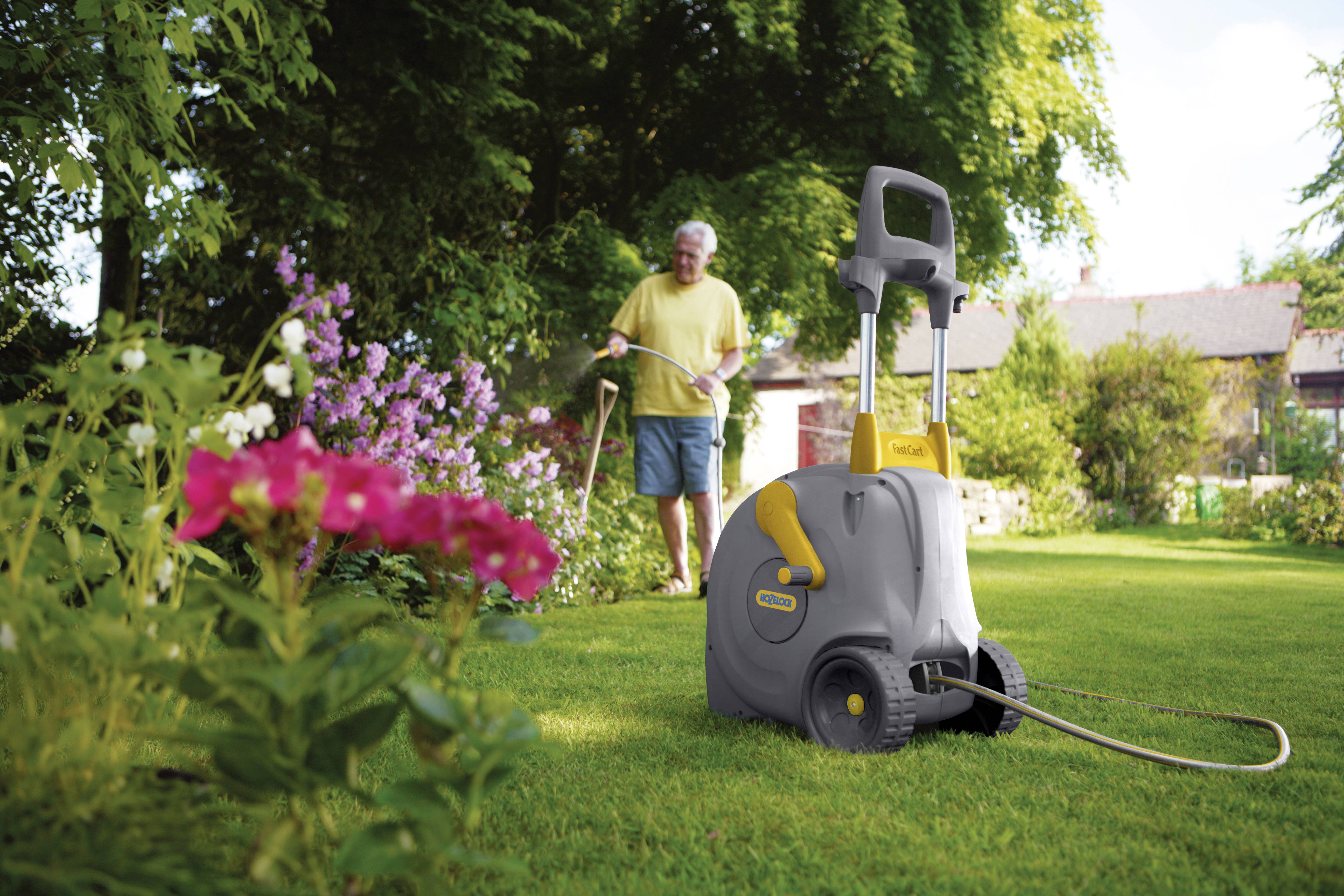 Een man besproeit een tuin met een waterslangenwagen op een zonnig gazon, omringd door bloeiende bloemen en bomen.