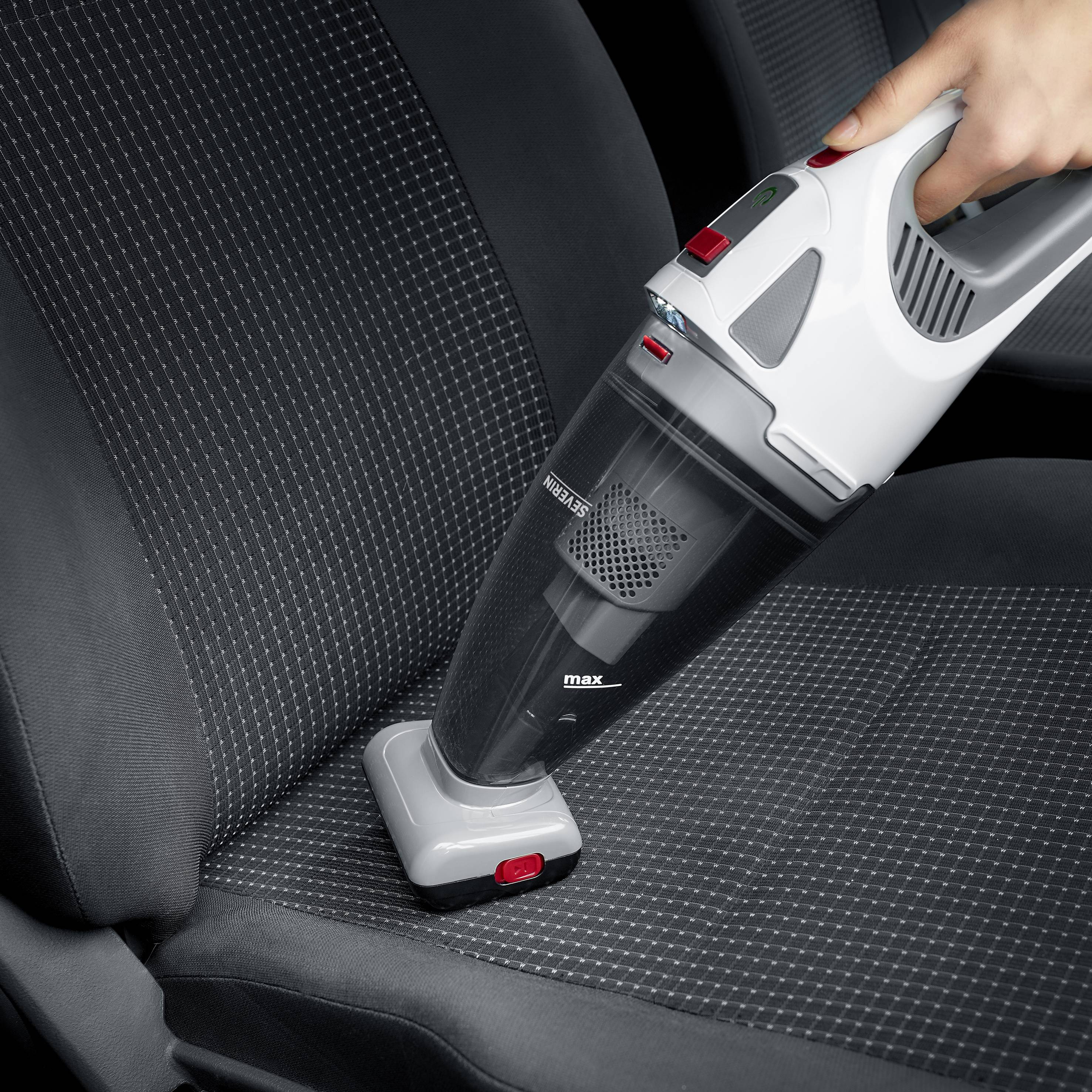 Een hand houdt een draagbare stofzuiger vast die de zitting van een auto schoonmaakt. De stofzuiger is zwart-grijs met een rode knop.