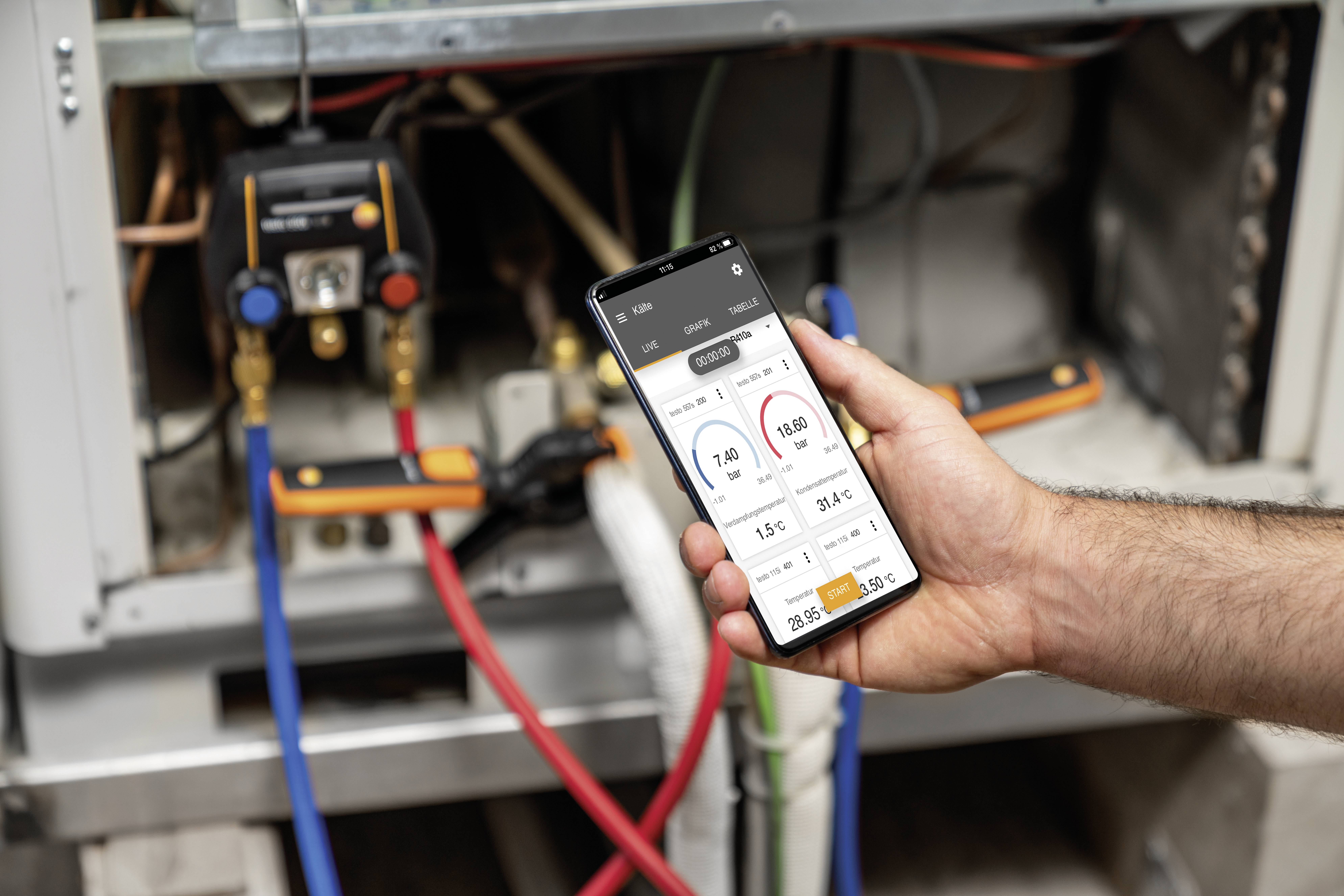 Een persoon houdt een smartphone voor een verwarmingsinstallatie. Op het scherm worden temperatuurgegevens en instellingen weergegeven.