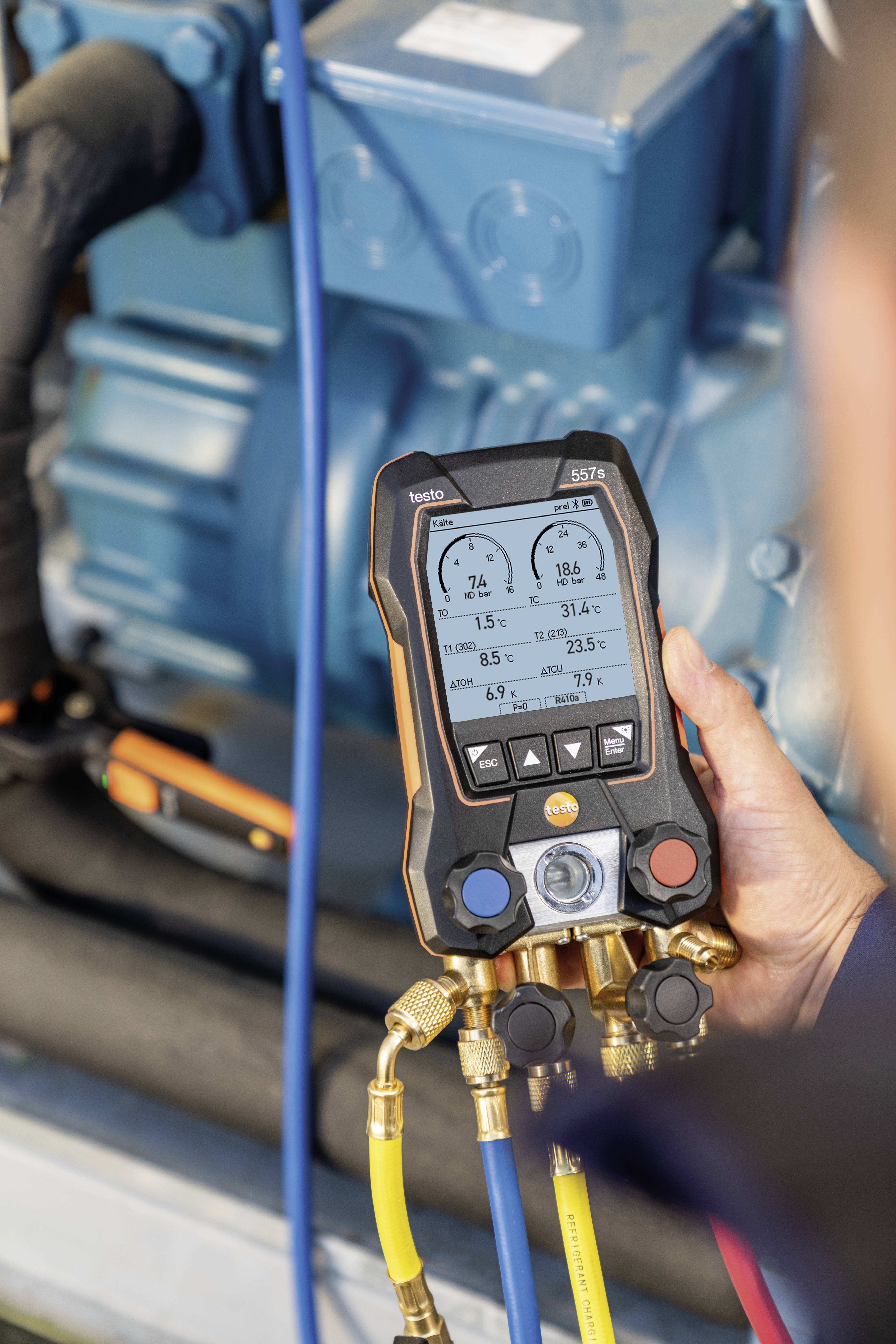 Een persoon houdt een digitaal meetinstrument vast dat druk- en temperatuurwaarden weergeeft voor een blauwe technische installatie.