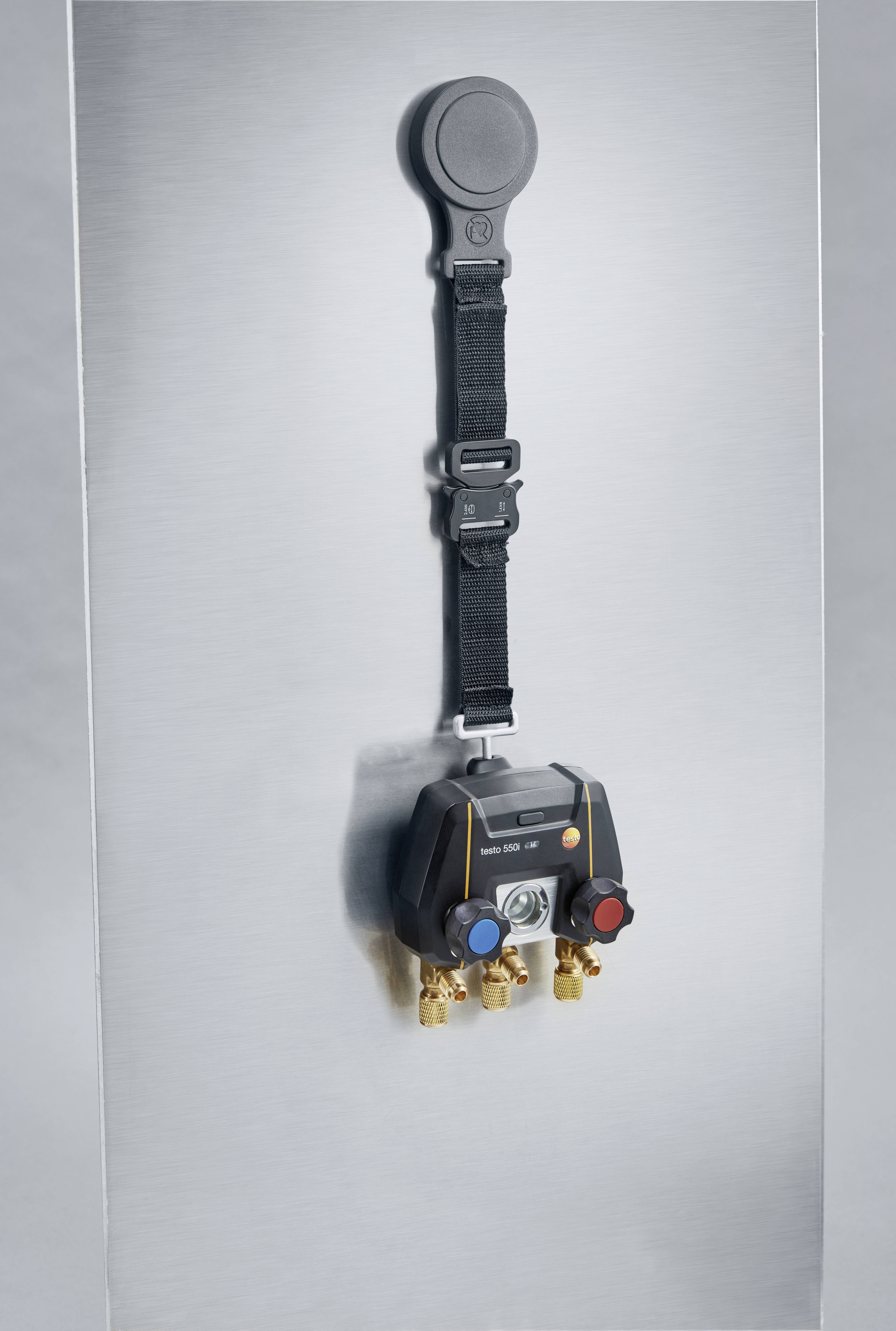 Een digitaal manometer met vier aansluitingen dat aan een metalen haak hangt. Het heeft kleurgecodes ventielen in rood en blauw.