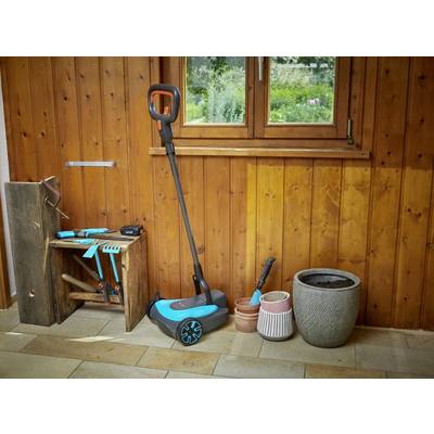Een draadloze bezem staat in een ruimte met houten wandbekleding. Ernaast liggen tuingereedschappen en enkele lege bloempotten.