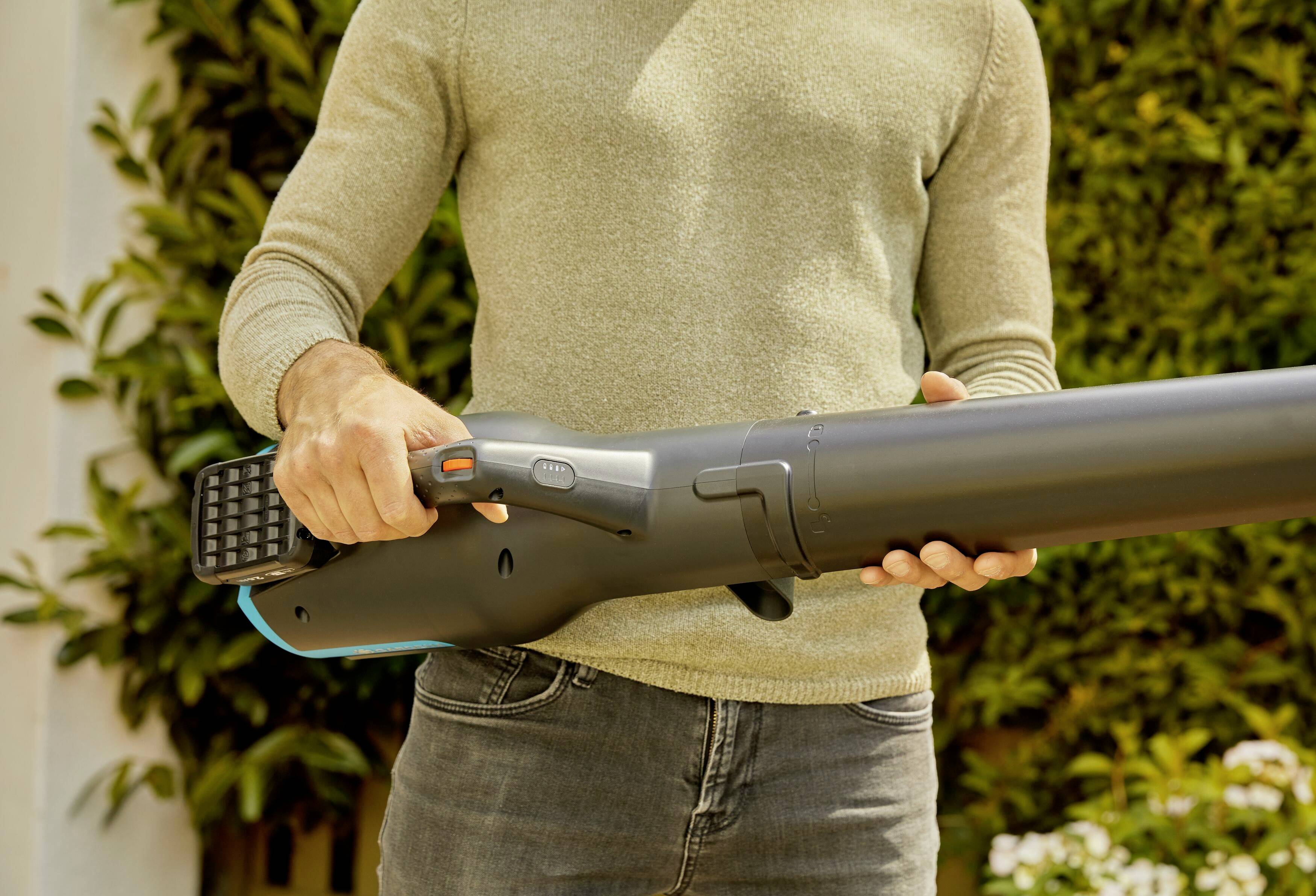 Een persoon houdt een groot, donker apparaat buiten vast. Het apparaat ziet eruit als een bladblazer en wordt met beide handen bediend.