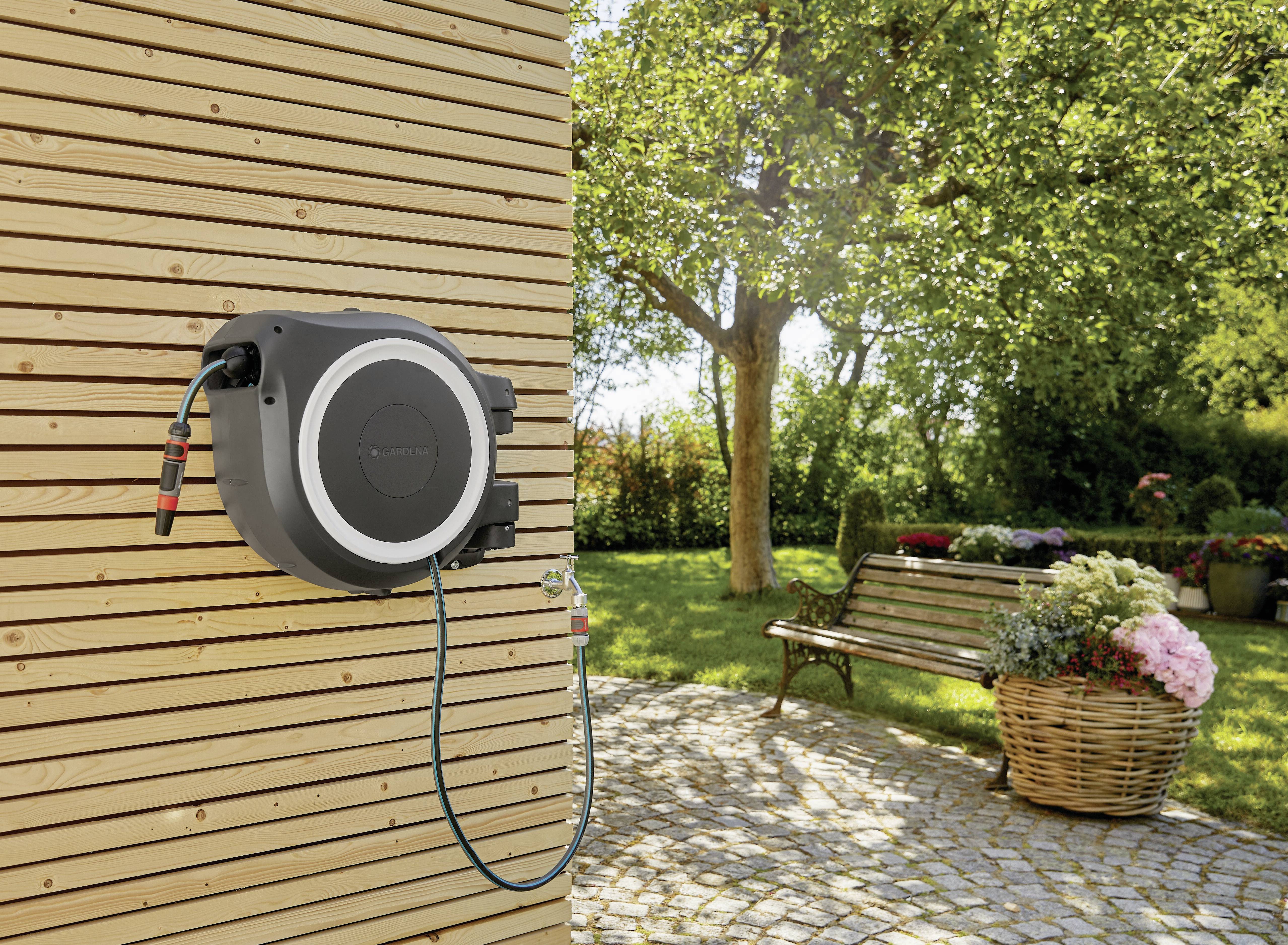 Een automatische tuinslangoproller is gemonteerd aan een houten muur. Op de achtergrond is een groene tuin met bomen en bloemen te zien.