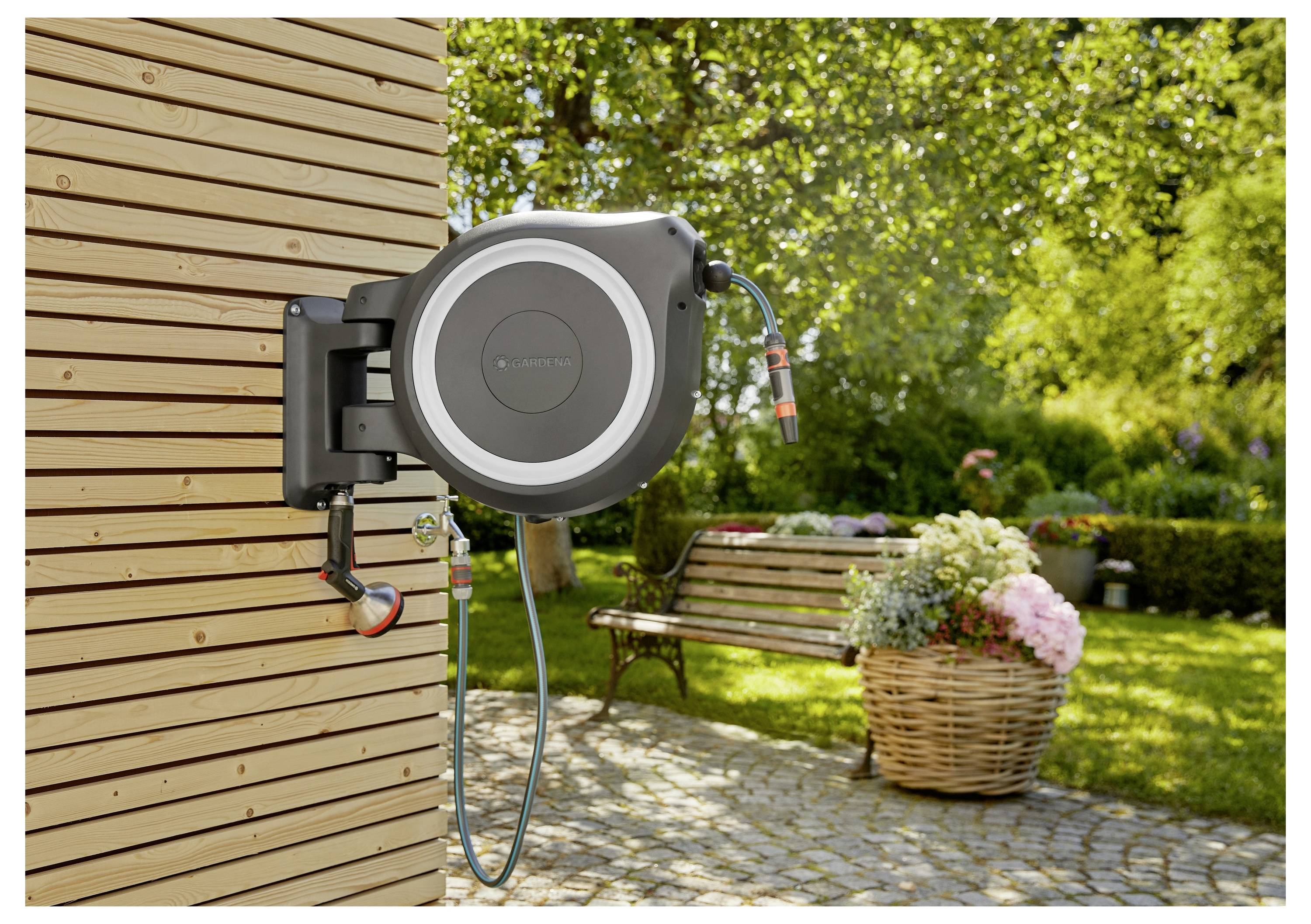 Een intrekbare tuinslang gemonteerd op een houten muur in een weelderige tuin. Een mand met bloemen en een bank bevinden zich op de achtergrond.