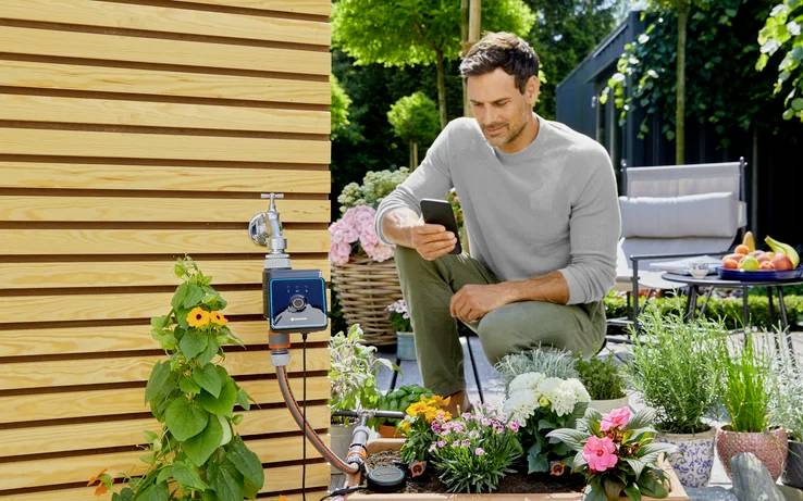 Een persoon in een tuinomgeving gebruikt een smartphone nabij bloeiende bloemen en een tuinslang die aan een houten muur is bevestigd. Gezellige zithoek op de achtergrond.