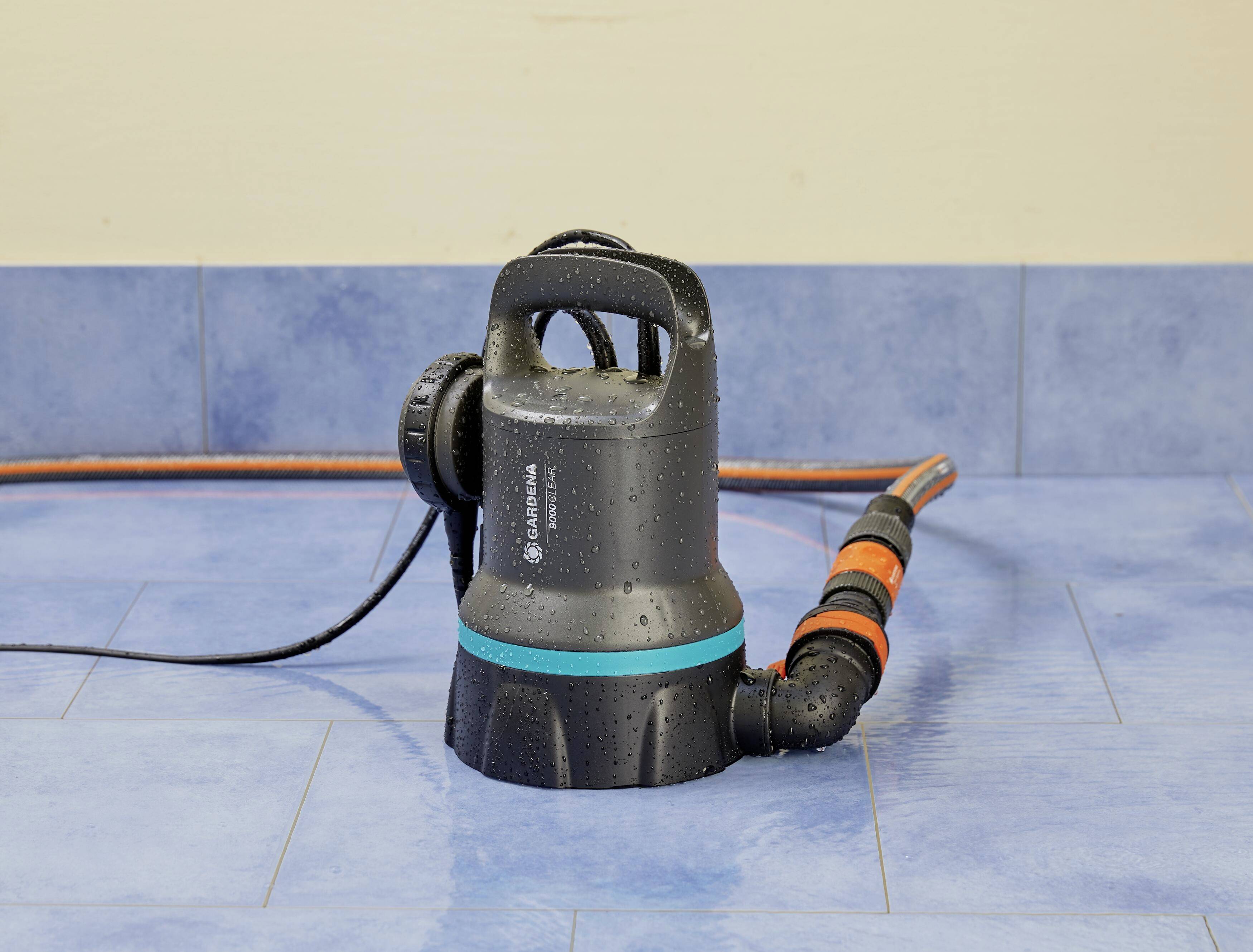 'Een zwarte waterpomp staat op blauwe vloertegels, met slangen die eraan bevestigd zijn. De pomp is nat van het water.'